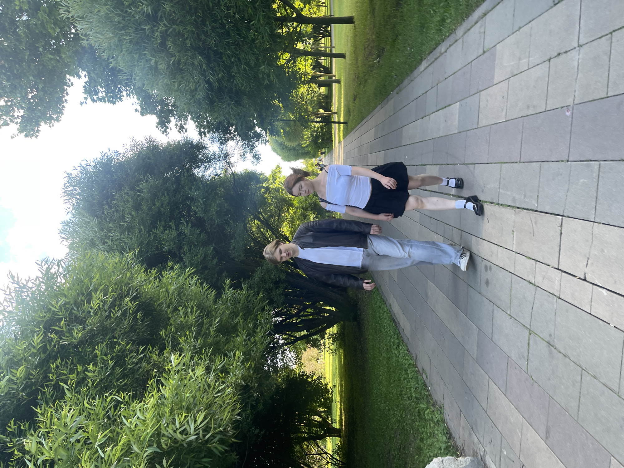 Two individuals, a young man and a young woman, are walking down a paved park pathway lined with trees. The man, on the left, wears a brown leather jacket over a white t-shirt and light-colored jeans. The woman, on the right, is in a black skirt and a white off-the-shoulder top. Both are walking towards the camera. The pathway is made of large, rectangular concrete tiles. Lush green grass borders the pathway on both sides, and mature trees with dense foliage form a canopy overhead, creating a dappled sunlight effect. The scene is set in a park, likely in Moscow, Russia, given the provided context, and the atmosphere appears serene and summery.