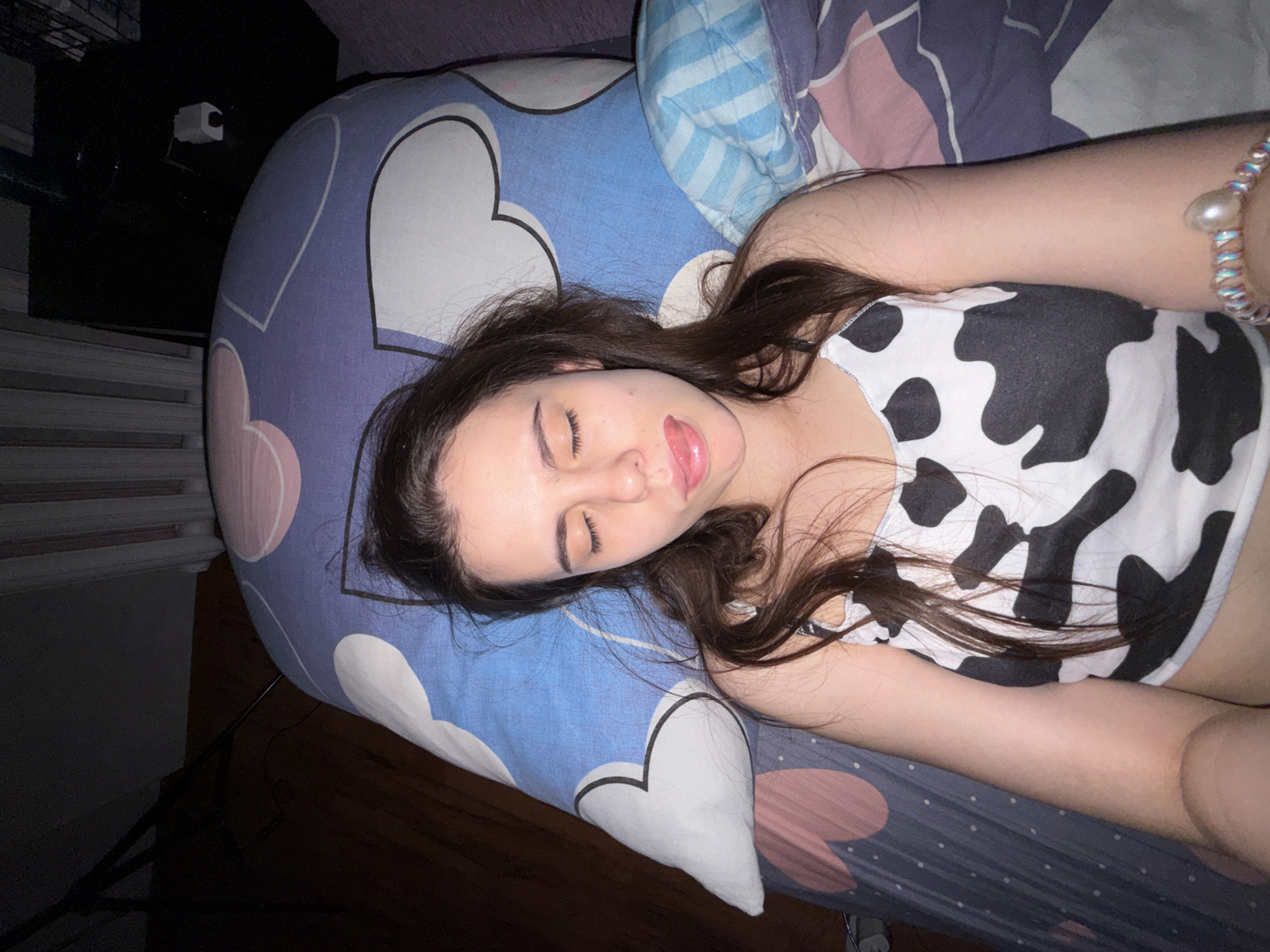 A young woman is lying on a bed. She is lying on her back, with her eyes closed and her tongue sticking out slightly. She is wearing a cow-print top and a bracelet on her right wrist. The bed has a heart-patterned pillow and a striped blanket. The scene is indoors, with a dark object in the upper left corner. The location and city are not discernible from the image.