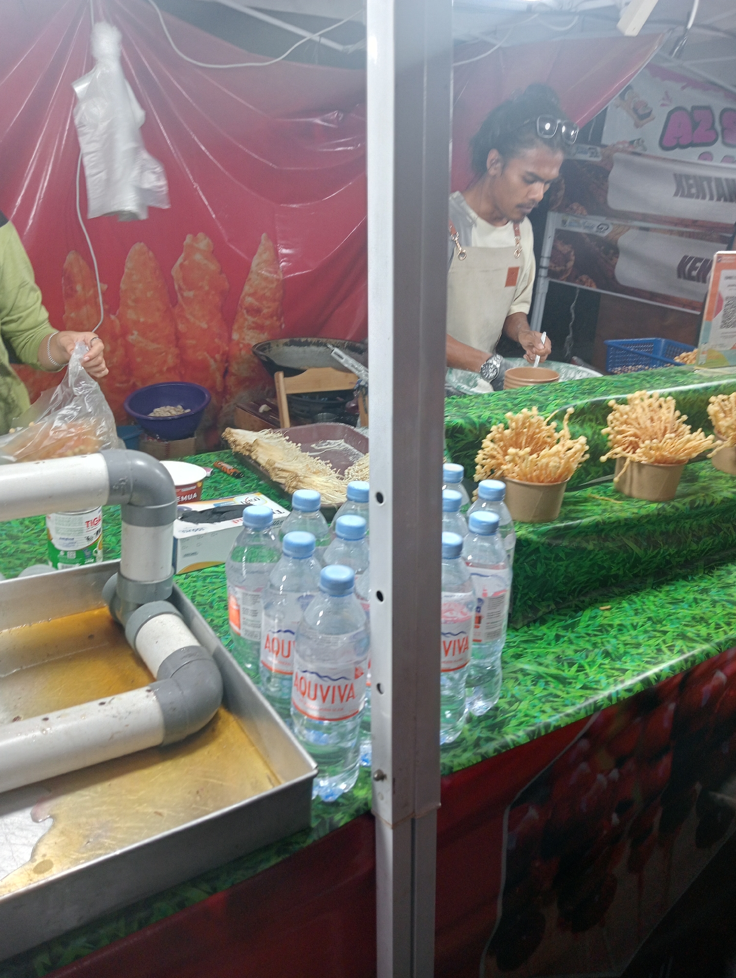 The image depicts a street food stall in Jambi, Indonesia, with a focus on prepared food items and beverages. In the foreground, a metal tray with a PVC pipe system contains a yellowish liquid, possibly oil or broth. Several plastic bottles of water, some labeled "AQUAVIVA," are arranged on a green, grass-patterned surface. Behind these, a stall worker with dark hair pulled back and sunglasses perched on his head is visible, wearing an apron and holding a utensil over a brown bowl. The background features a red tarp, decorative food imagery resembling fried items, and signage that includes the partial text "AZ S" and "KENTANG." A blue basket is also visible, containing small objects. The overall scene suggests a bustling food stall operation.