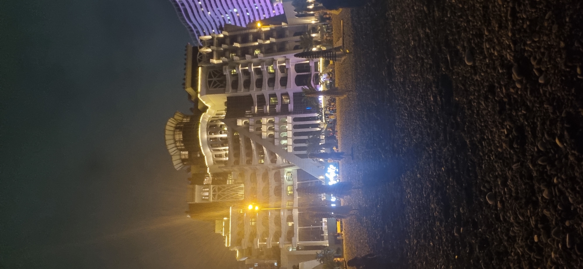 This image depicts a nighttime scene on a pebble beach in Batumi, Georgia, with a large, illuminated hotel complex as the dominant background element. The hotel, characterized by its terraced architecture and extensive exterior lighting, appears to be a prominent landmark. Several beach umbrellas are visible on the shore, some appearing closed and others partially open, suggesting preparation for or the end of a beach day. A few individuals are scattered on the beach, some appearing to be walking or standing, their forms silhouetted against the illuminated hotel. The sky is dark, indicating it is nighttime. The foreground is occupied by the pebble-strewn beach, with the stones clearly visible due to the artificial lighting. The overall impression is of a vibrant urban beach resort at night.