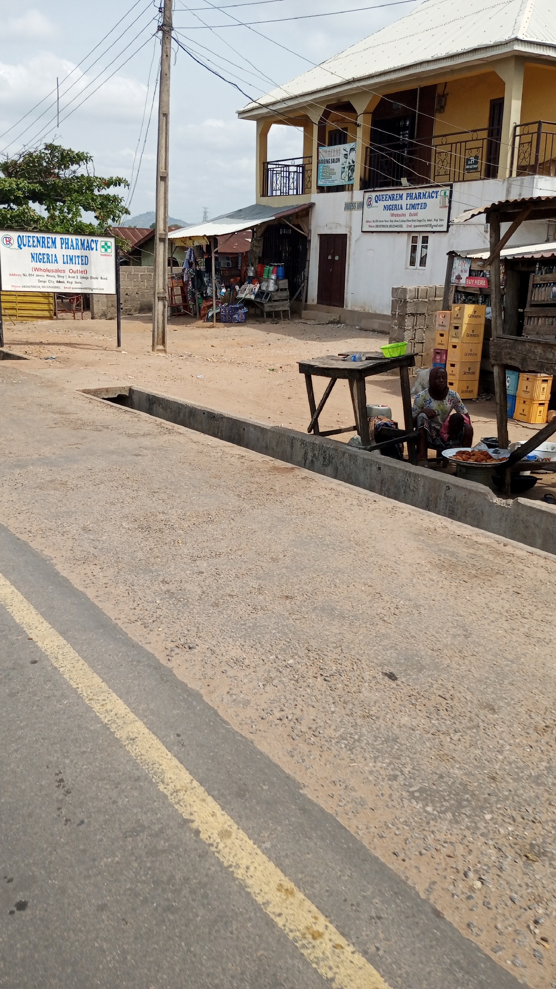 The image depicts a street scene in Zango-ndaji, Nigeria. A woman is seated next to a table and cooking equipment, preparing food for sale. The scene is set near a concrete drainage ditch, and a road with a yellow dividing line is visible in the foreground. A sign for "QUEENREM PHARMACY NIGERIA LIMITED" is prominent in the background. The building hosting the pharmacy also features a "BARBING SALON" and several vending stores. Stacks of Guinness beverage crates are present on the right side of the image. The area appears to be a commercial roadside.