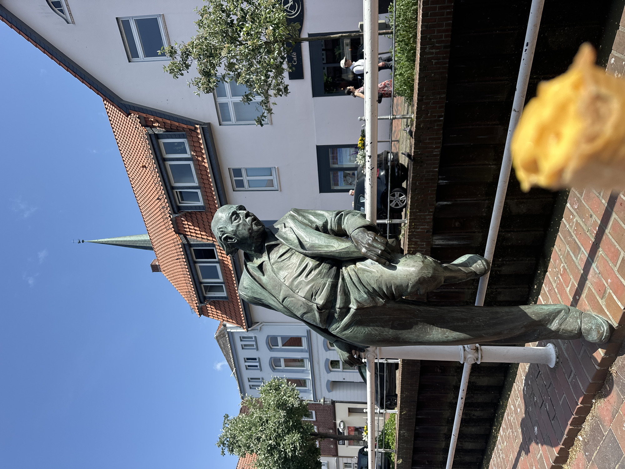 Here is a description of the image:
The image shows a bronze statue of the German writer Erich Kästner leaning against a white metal railing in Buxtehude, Germany. The statue depicts Kästner in a suit, holding a book. Behind the statue are buildings with red tile roofs and white facades. A cafe is visible in the background. A blurry object is in the lower right foreground, possibly food. The sky is blue, and there are trees nearby. The setting is a brick walkway along a canal.