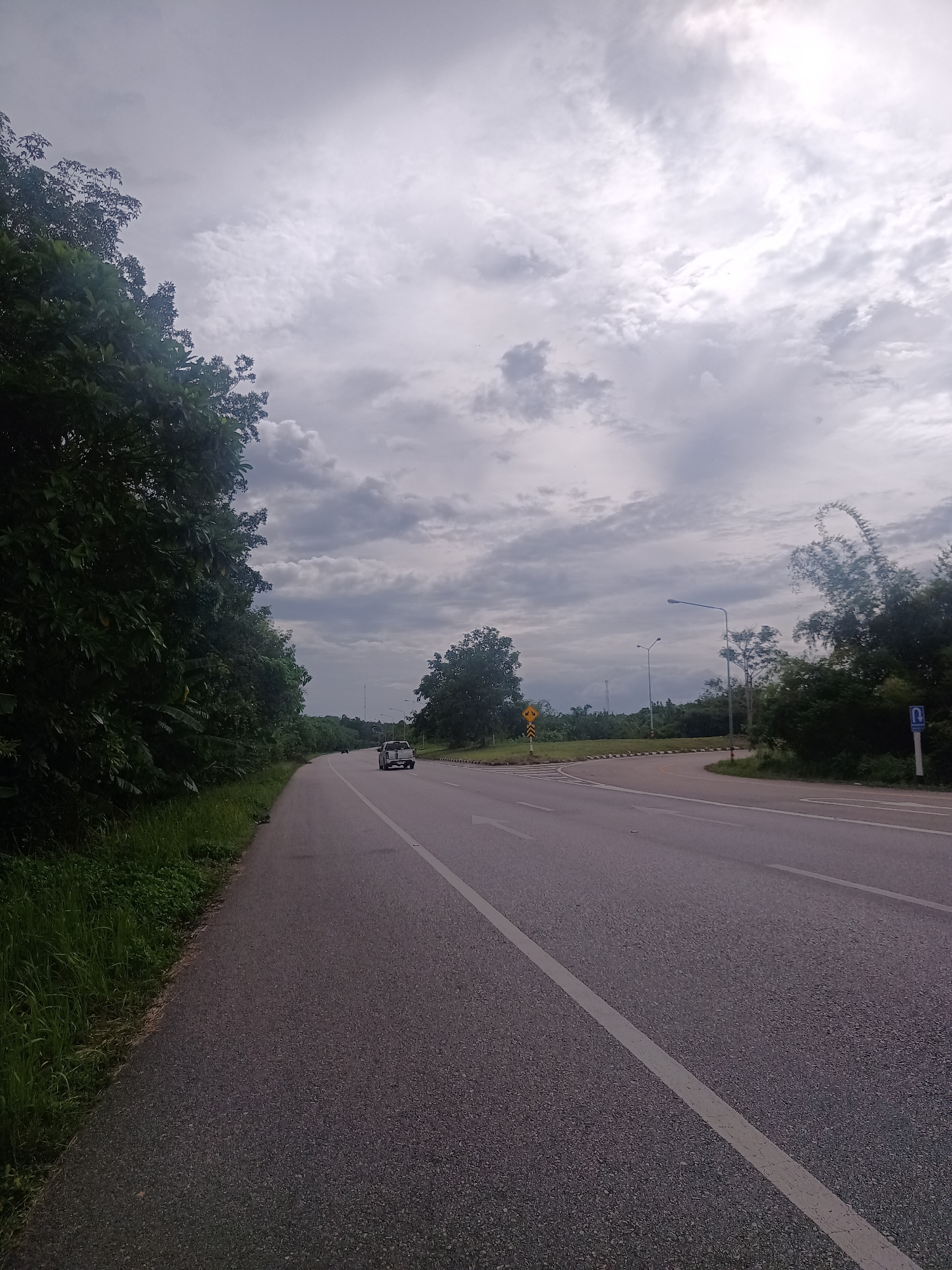 The image depicts a stretch of road in Khian Sa, Thailand under an overcast sky. The road, composed of asphalt, has a white lane marker on the right side and white directional arrows indicating straight ahead. A car is visible on the road in the distance. Dense green foliage lines the left side of the road, while the right side reveals more open vegetation and trees. Street lights are visible along the road. A yellow traffic sign and a blue parking sign are present on the right side of the road.