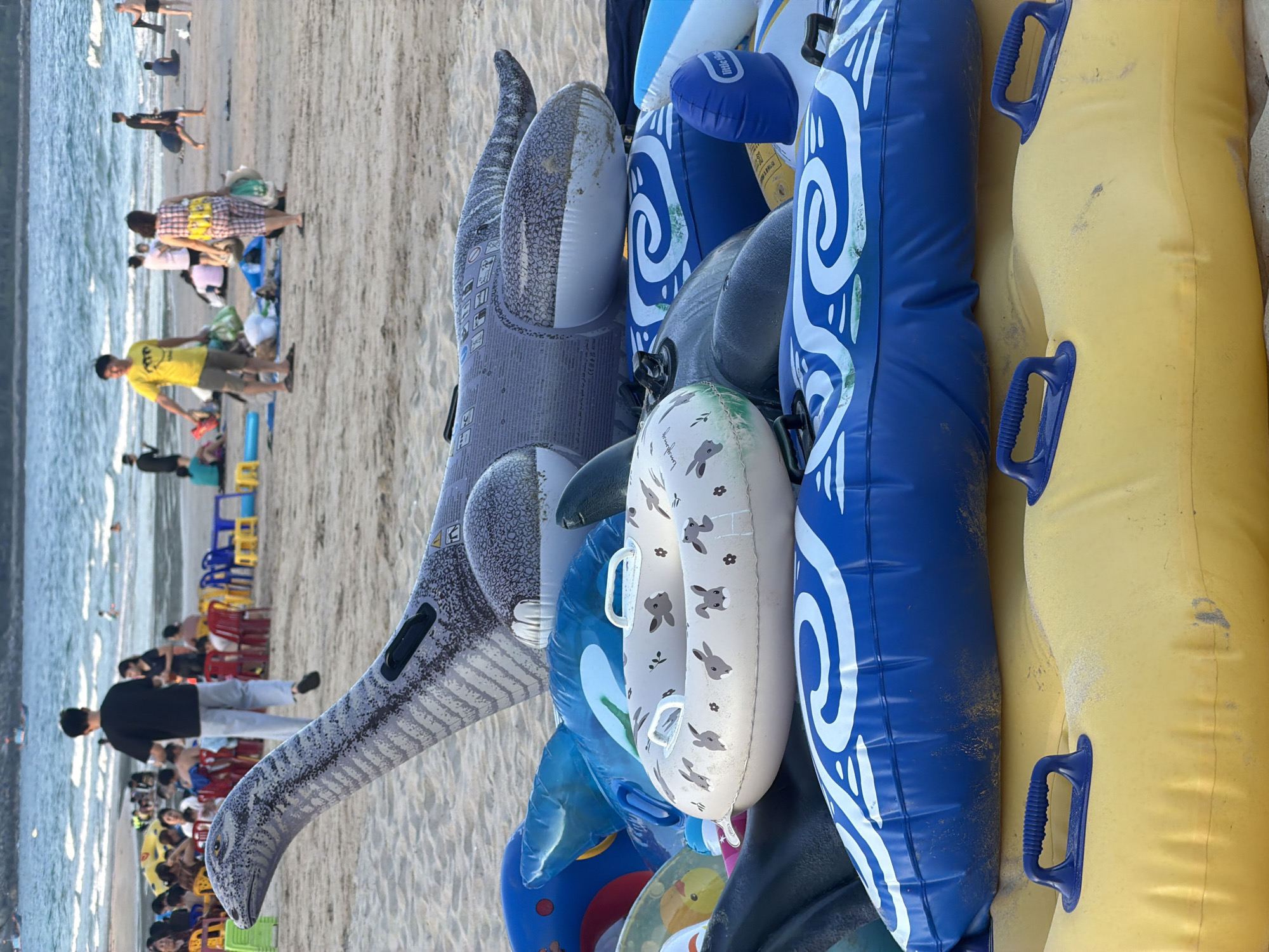 In Cam Lam, Vietnam, a pile of inflatable pool toys rests on a sandy beach, with the ocean and a few distant beachgoers visible in the background. The foreground is dominated by the colorful inflatables, including a large, gray dinosaur-shaped float, several blue and white swirl-patterned tubes, a light-colored tube with rabbit graphics, and a yellow tube with blue handles. Some of the inflatables appear worn and dusty from use. In the middle ground, people are scattered on the sand, some sitting at tables under umbrellas, while others are walking or standing near the water. The sea is calm with gentle waves.