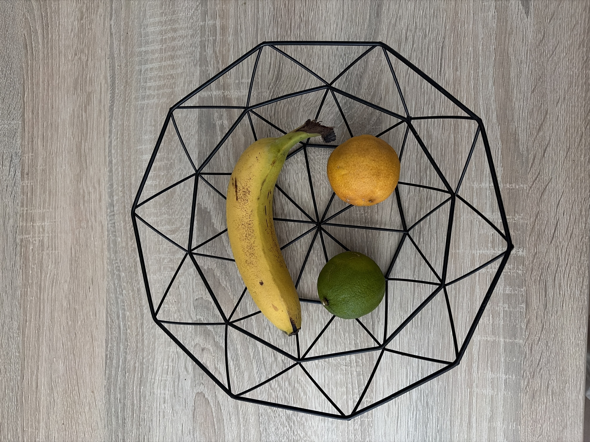 A geometric black wire fruit basket contains a yellow banana, a green lime, and an orange. The basket sits on a light-colored wooden surface. The scene is viewed from directly above.