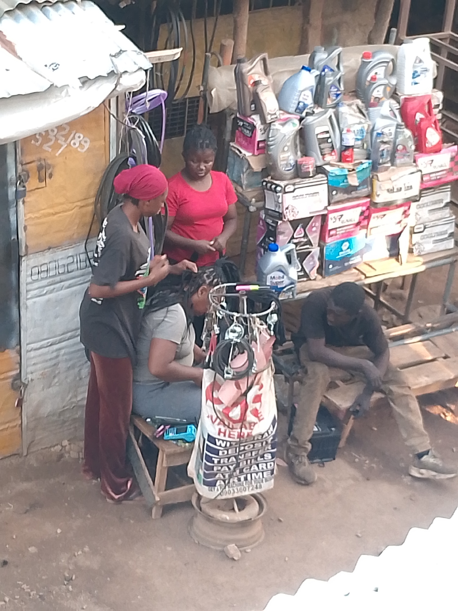 The image depicts an outdoor scene in Bukuru, Nigeria. Two women are braiding the hair of a seated woman near a makeshift storefront with a corrugated metal exterior. A third woman stands nearby observing. A man sits on a bench next to a display of motor oil and car batteries. Cables and parts are visible in the background. The storefront displays hand-painted signs and phone numbers.