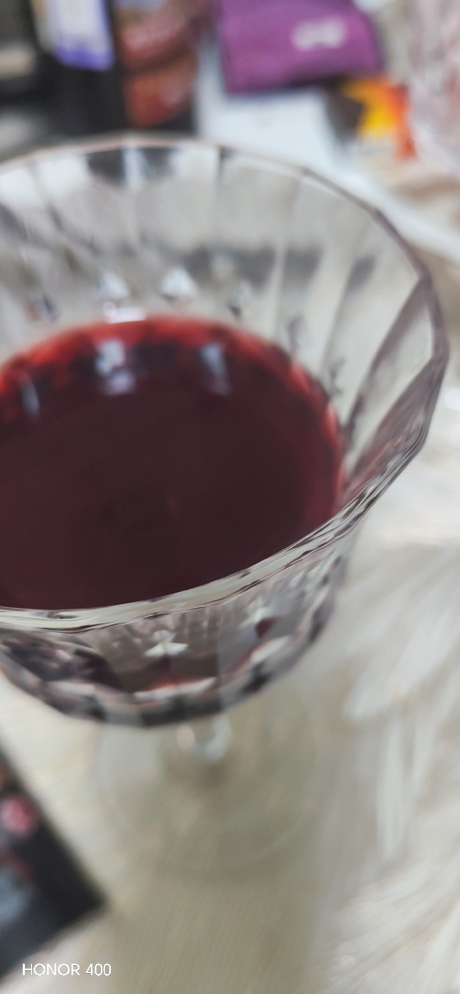 A close-up, high-angle photograph captures a clear, faceted glass, partially filled with a dark red liquid. The liquid's surface reflects ambient light. The glass is positioned over a light-colored, textured surface, resembling white faux fur or a thick fabric. The background is intentionally blurred, displaying indistinct shapes including a dark bottle, a purple object, and other unidentifiable items, suggesting an indoor environment. No individuals are present within the frame. A "HONOR 400" watermark is visible in the lower left corner of the image. The scene is located in the city of Noviy, Russia.