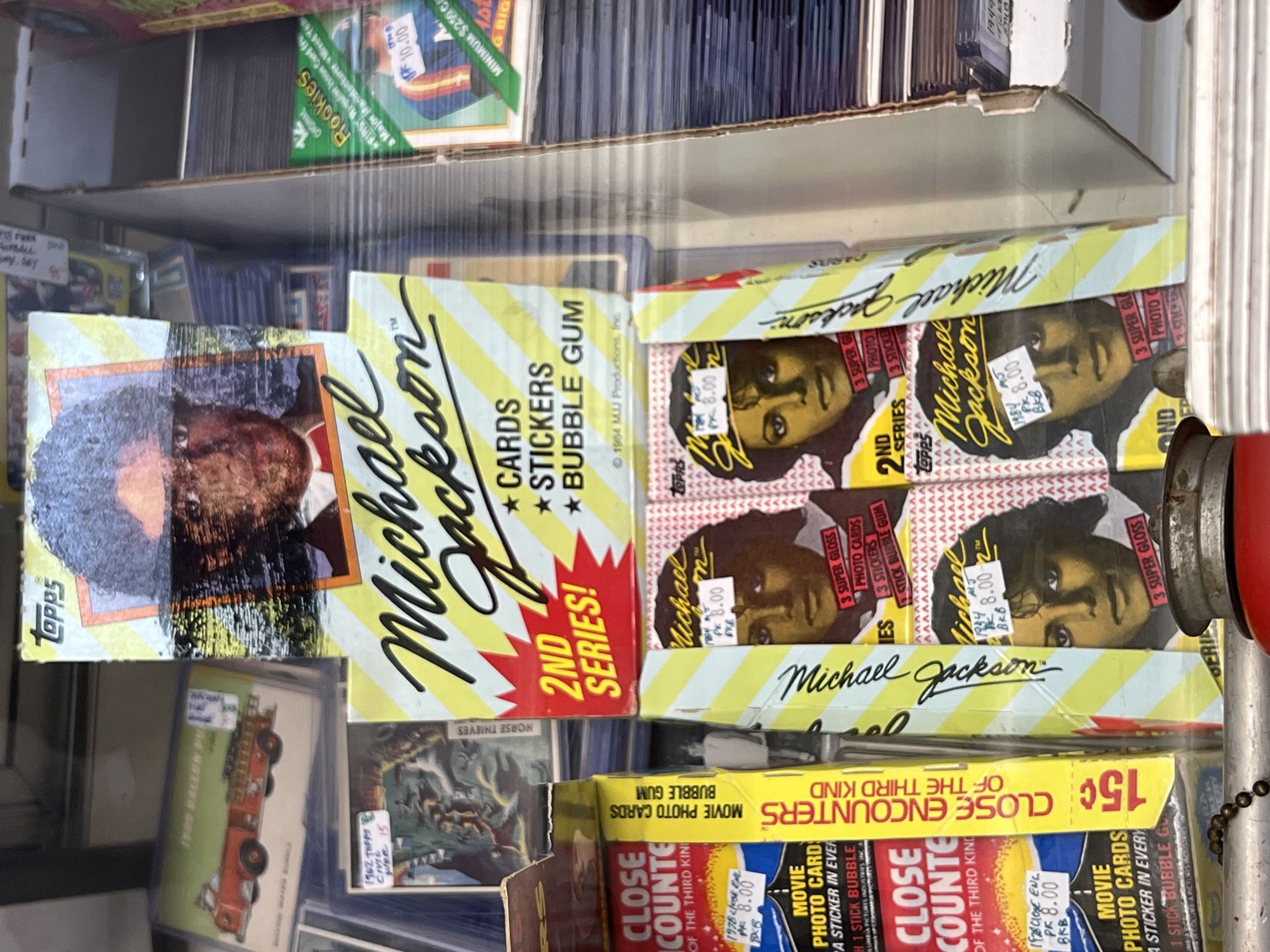 The image shows a shelf in a shop, likely in City of Newburgh, United States of America, filled with vintage trading card packs. Several packs of "Michael Jackson 2nd Series" cards by Topps are prominently displayed. The price "8.00" is written on the packs. Also visible are packs of "Close Encounters of the Third Kind" movie photo cards and older baseball cards. The packs are stacked and arranged in rows, creating a cluttered but organized appearance.