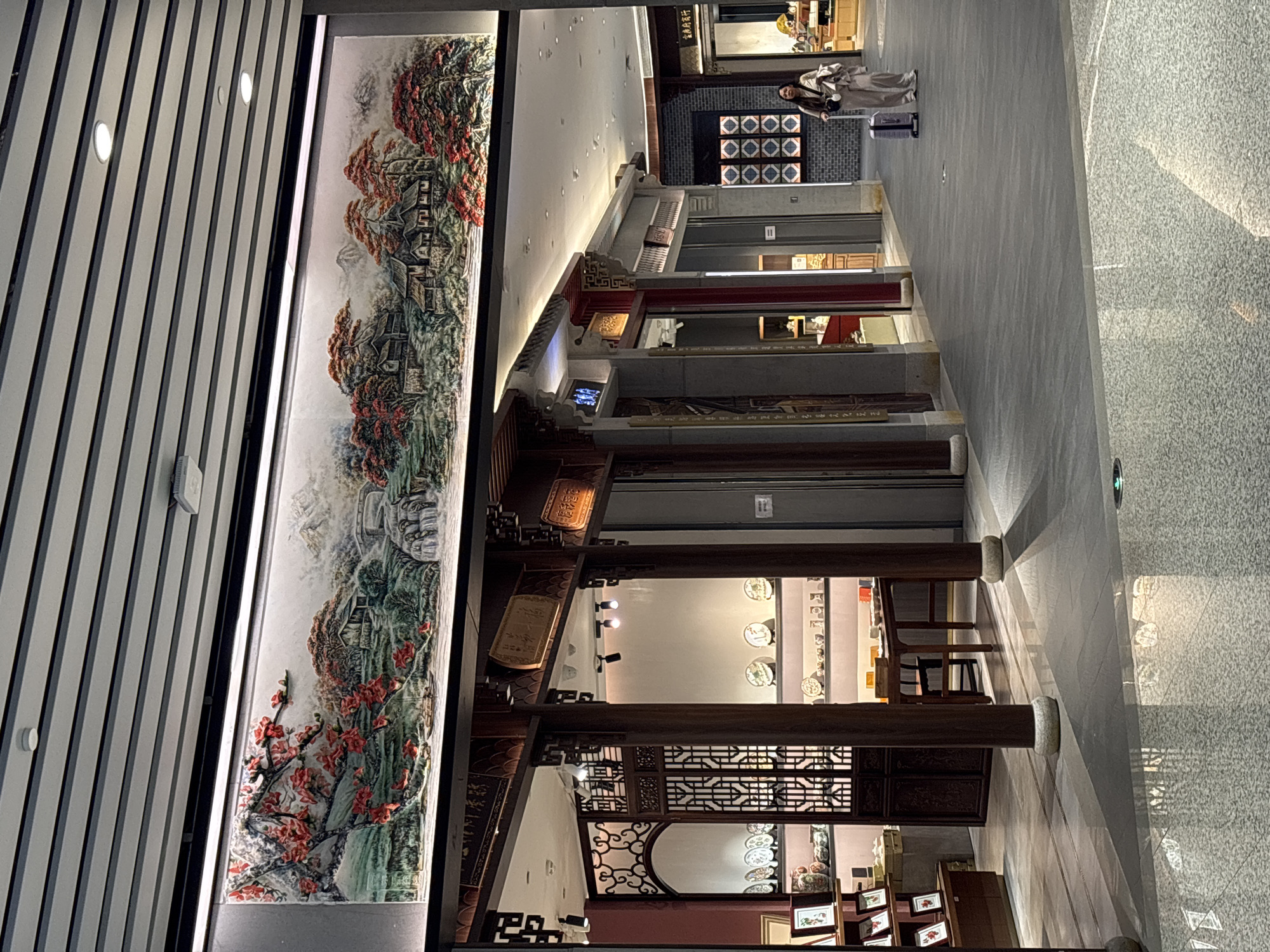An indoor shot of a museum or exhibit in the city of Khimki, Russia. A mural with red and green vegetation and buildings is visible on a wall. Wooden beams extend into the room. Plates are visible on the wall. A person is standing by the wall. Chairs and tables are visible. Other displays are present.