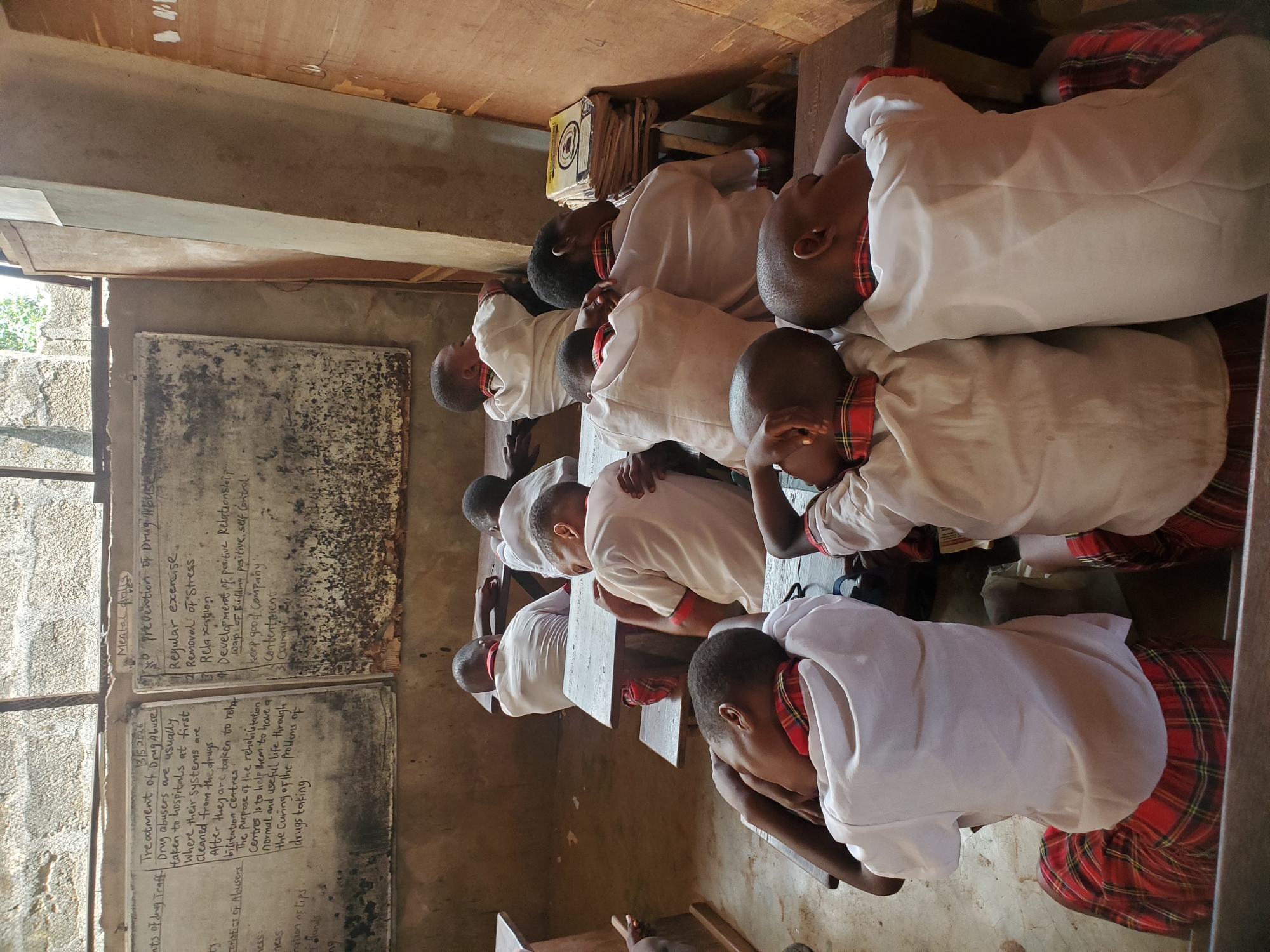 A classroom in Ukwu-Abwa, Nigeria, features rows of students in white shirts and plaid skirts/trousers resting their heads on their desks, possibly sleeping. The classroom has a blackboard on the left filled with notes about drug abuse and its treatment, indicating a health or educational setting. The environment appears to be a basic structure, with simple wooden desks and chairs. There are papers and notebooks stacked on a shelf above the desks. The room is lit by natural light coming through a window on the left.