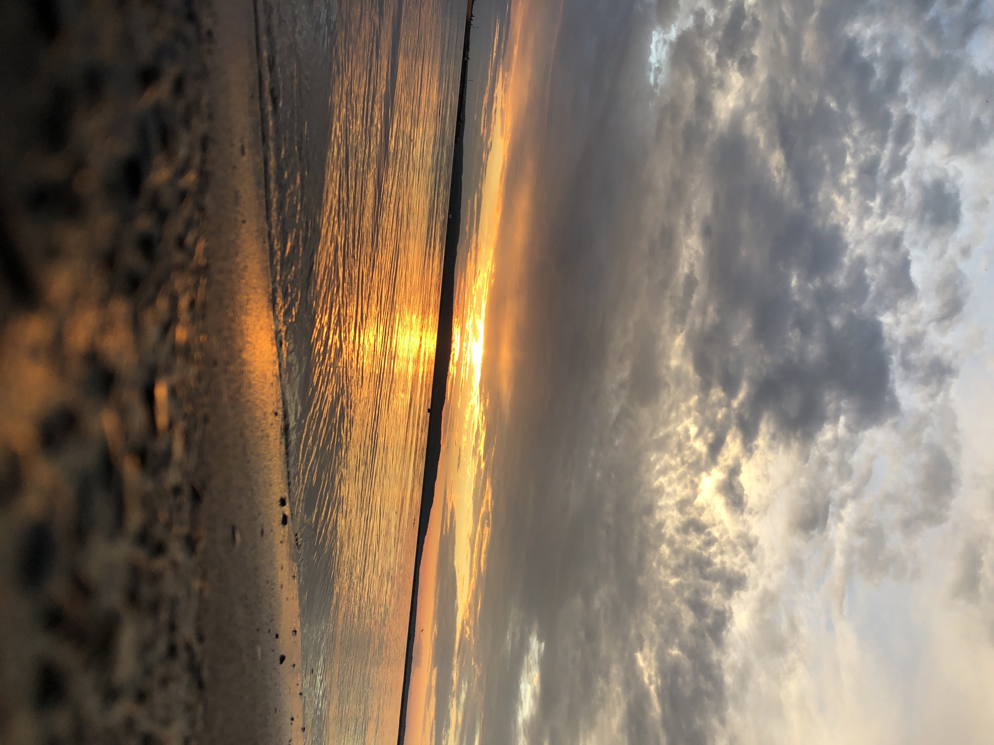 The image depicts a serene sunset over the Kama River in Chaikovsky, Russia. The sky is filled with dramatic, cloudy formations, with streaks of orange and yellow light peeking through as the sun dips below the horizon. The water reflects the vibrant colors of the sunset, creating a shimmering, golden pathway across the surface. The foreground shows a sandy beach with scattered pebbles, leading down to the gentle lapping waves. The overall scene is tranquil and picturesque, showcasing the natural beauty of the Chaikovsky landscape during twilight.
