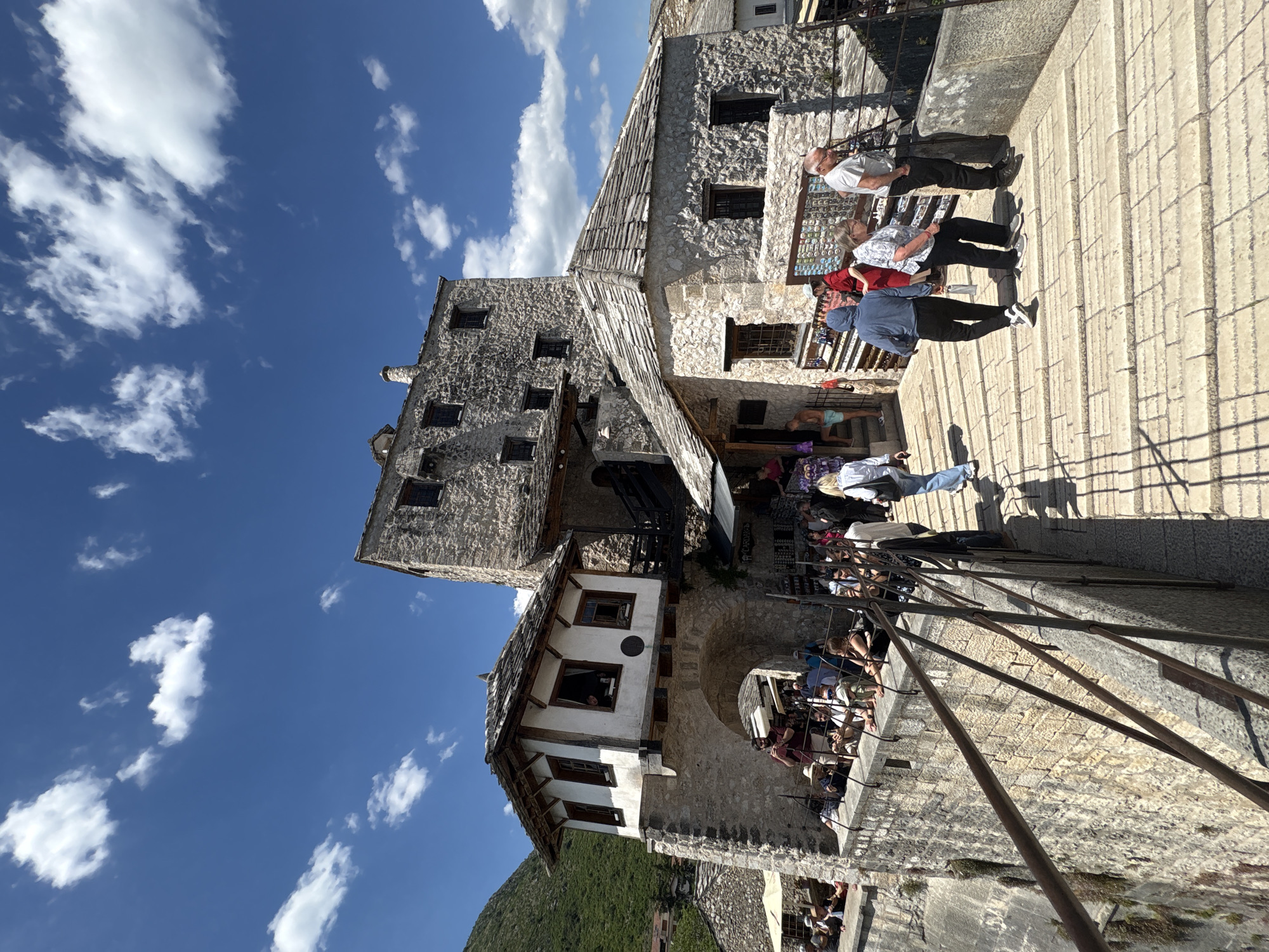 This photo depicts a view of a stone building, likely a historic structure, in Mostar, Bosnia and Herzegovina. The building has a light-colored stone facade, multiple windows, and a tiled roof. The architecture appears to be of traditional Ottoman style.
A group of people is positioned near the building, possibly tourists or locals interacting with the scene. They are walking, standing, or looking at displays of goods.
The sky is a bright blue with scattered white clouds. In the background, green vegetation suggests a natural landscape element. The camera angle is somewhat tilted, providing a dynamic perspective of the scene. Metal railings are visible in the foreground, hinting at a bridge or elevated walkway.