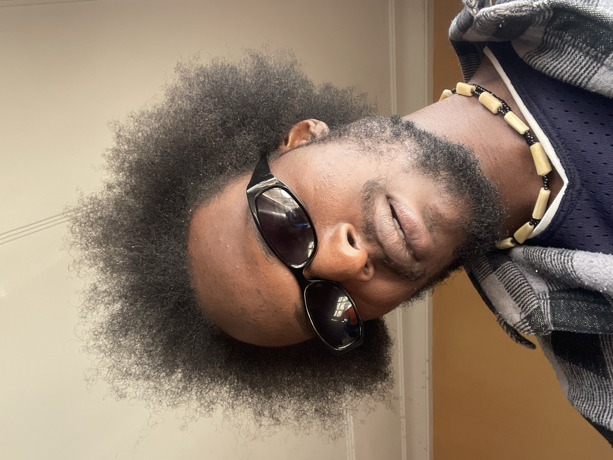 This is a close-up, selfie-style portrait of a Black man with a large afro hairstyle, wearing black sunglasses, a plaid shirt over a dark athletic jersey, and a necklace made of wooden beads and black seed beads. He has a beard and a goatee. The background is a plain, light-colored wall with a beige/orange tone on the right side, and a white ceiling with subtle molding visible at the top. There is a hint of natural light coming from the right. The man's head is tilted slightly to his left. The image captures his face and upper torso.
The provided image does not contain any discernible landmarks or clues that would allow for the specific identification of the location within the city of Jos, Nigeria.