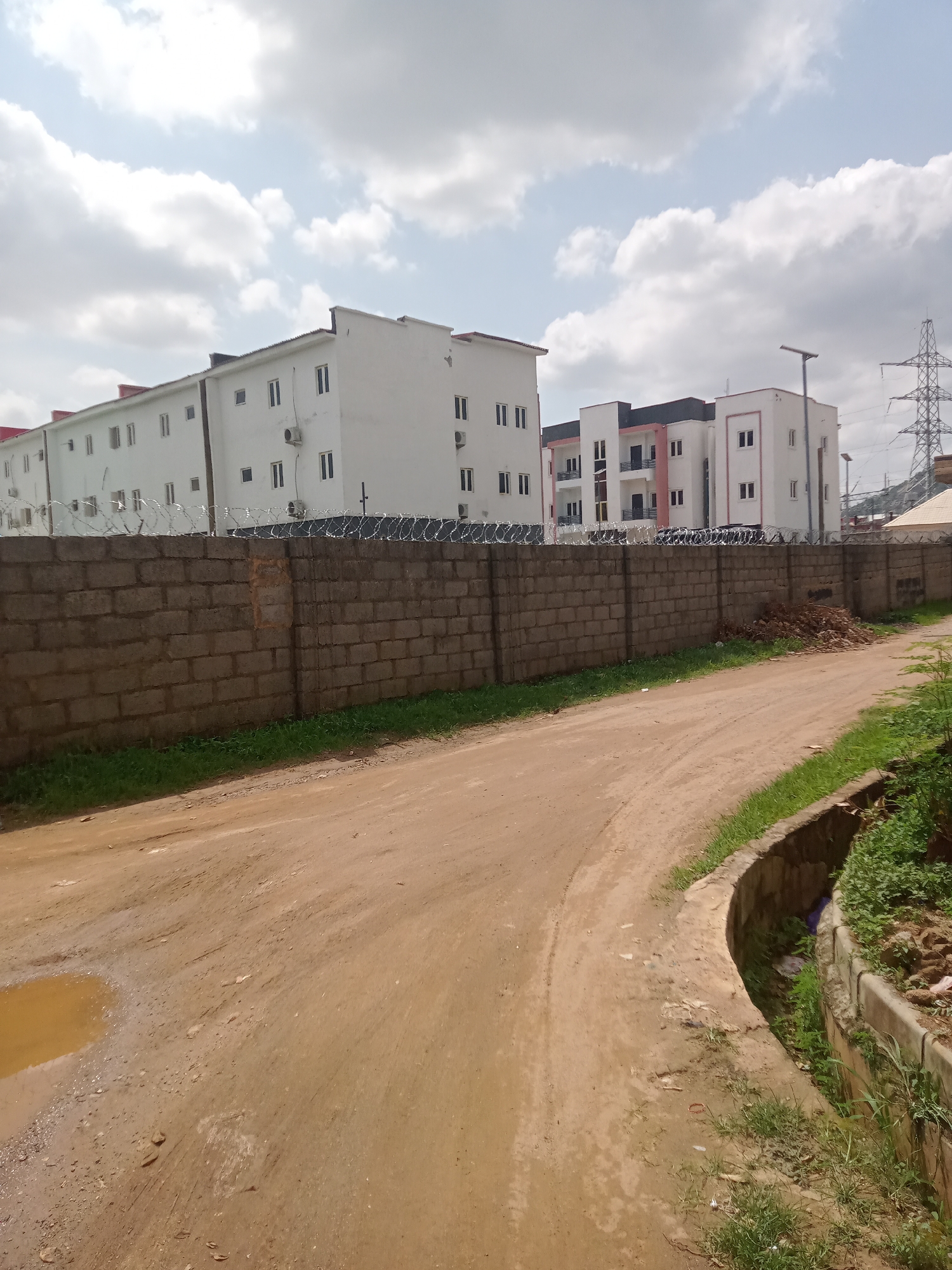 The image depicts a dirt road in Gwarinpa, Nigeria. A puddle of water is visible on the left side of the road. To the right of the road is a concrete drainage ditch with vegetation growing inside and along its edges. On the left side of the road is a long brick wall topped with barbed wire. Behind the wall are several white buildings with multiple windows. Power lines are visible in the background to the right. The sky is overcast with white and grey clouds.
