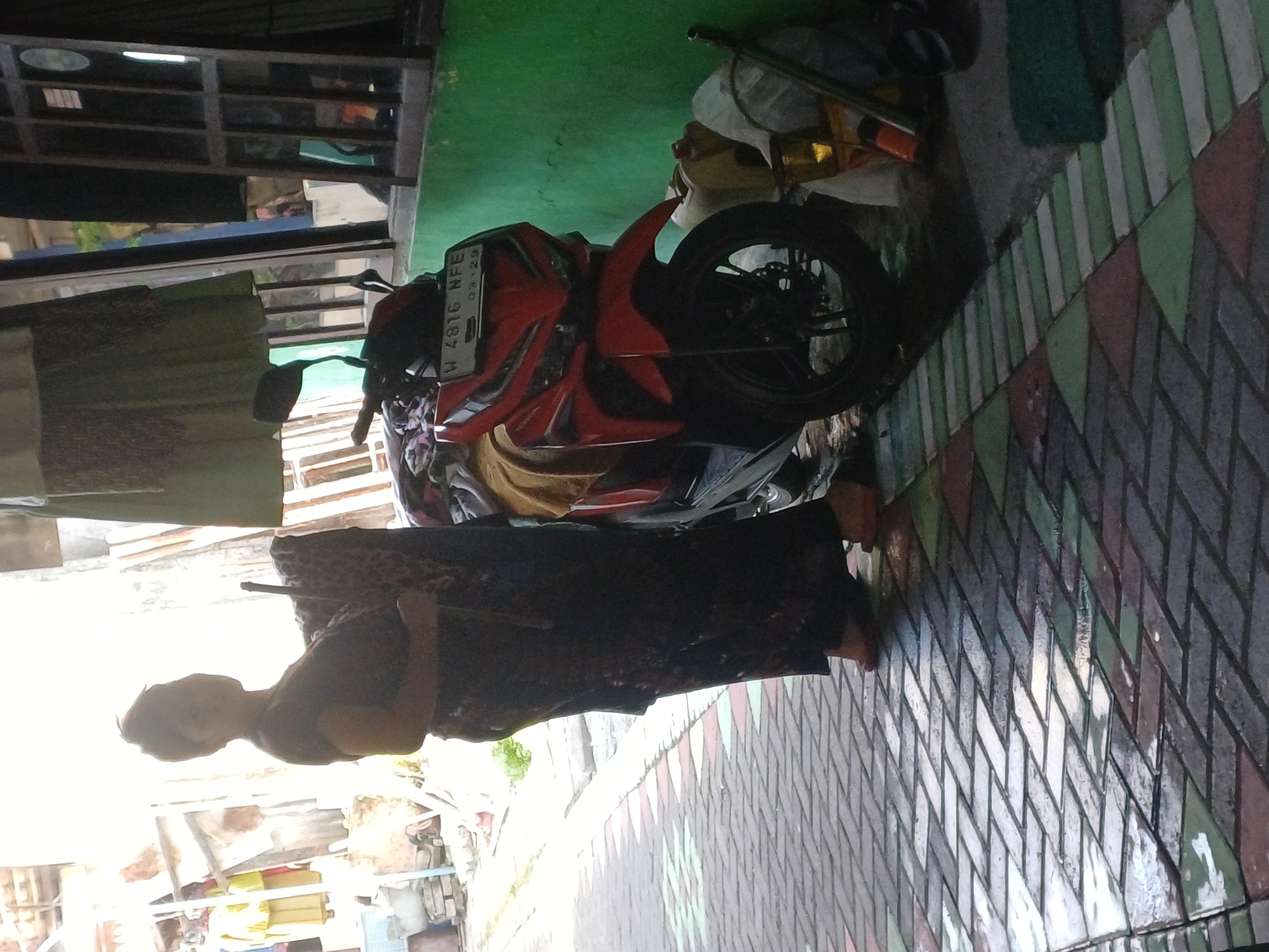 A person stands next to a red motorcycle in what appears to be an outdoor alleyway or walkway in Pulungan, Indonesia. The person, wearing a dark patterned dress and holding a patterned cloth, is positioned to the left of the motorcycle. The motorcycle, a red scooter with a license plate that reads "W 4816 NFE," is parked on a cobblestone path. The environment is characterized by a green wall on the right, a window frame, and hanging fabric to the left. The ground is paved with interlocking rectangular and triangular stones in various shades of gray, white, and green, with some areas appearing wet, suggesting recent rain. The lighting is bright, with the sun creating a strong contrast and overexposure in the upper left portion of the image. Various objects, including what appear to be containers and a basket, are visible near the motorcycle and along the green wall.