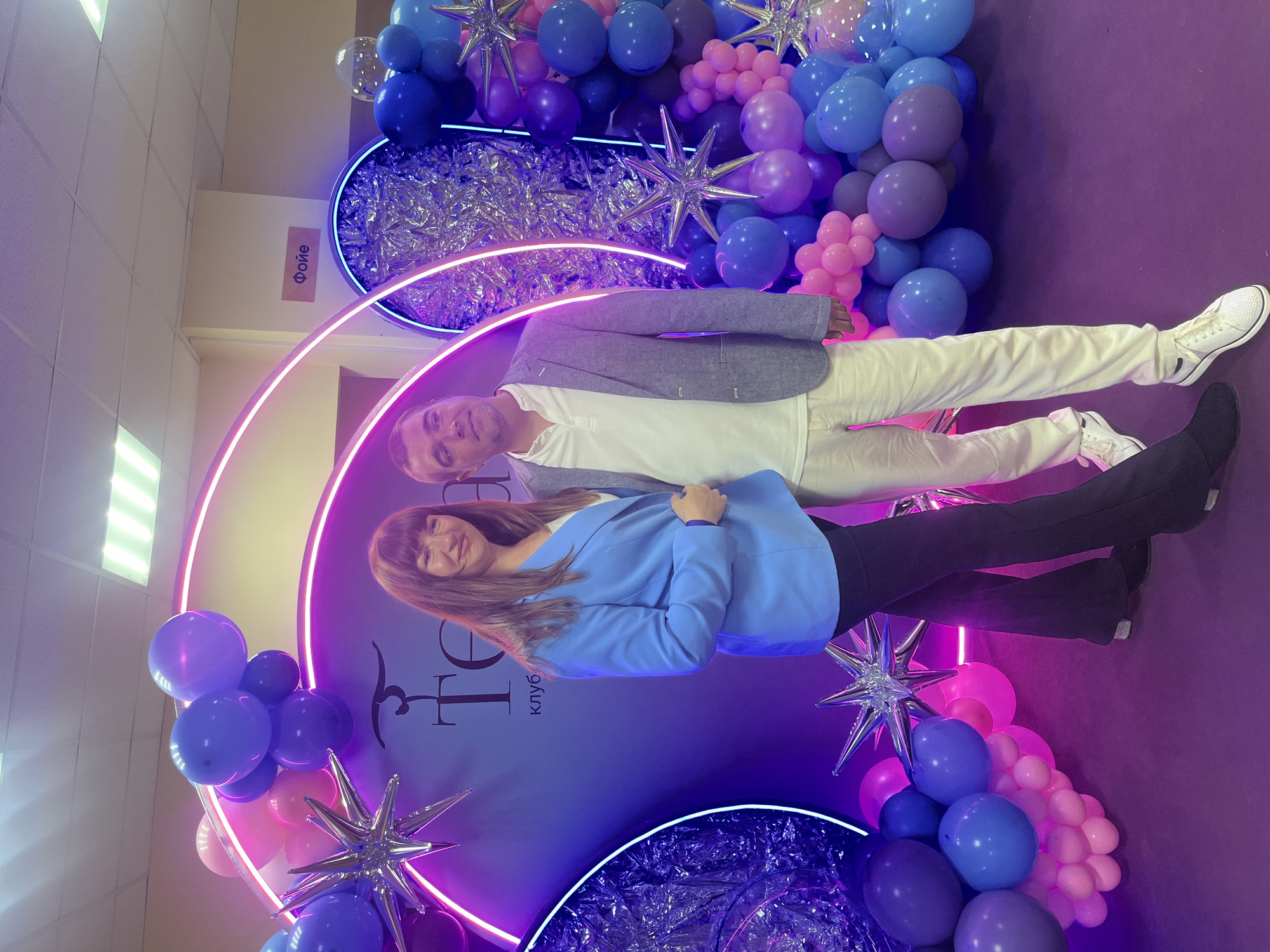 In Strubkovo, Russia, a man and a woman pose in front of a promotional backdrop. The man is wearing a gray blazer, white shirt, and white pants with white sneakers. The woman is wearing a blue blazer and dark pants with black shoes. The backdrop is a circle with the word "FEER клуь" written on it. The scene is decorated with purple and blue balloons and star-shaped ornaments. Neon lights frame the backdrop.
