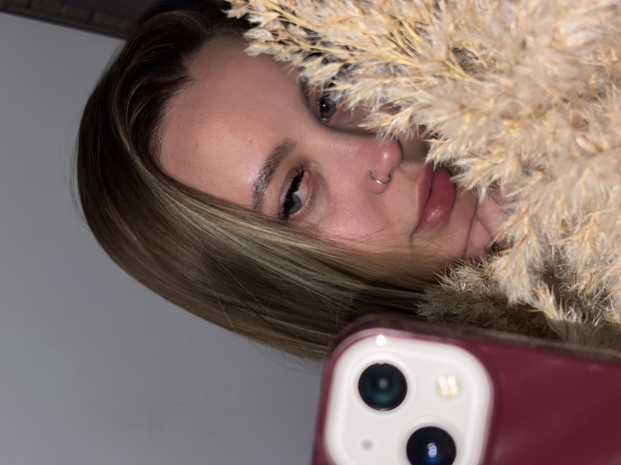 A young, light-skinned woman with blonde-streaked light brown hair is visible from the left side of her face to the center, and from her forehead to her lips. She has light grey-green eyes, a nose ring, and pinkish lips. She is holding a bundle of dried grasses or reeds, which are partially obscuring her face. The woman is holding a maroon-colored smartphone with a dual-lens camera in front of her. The background is a muted grey wall. The location is not identifiable as Port-Katon, Russia based on the image alone.