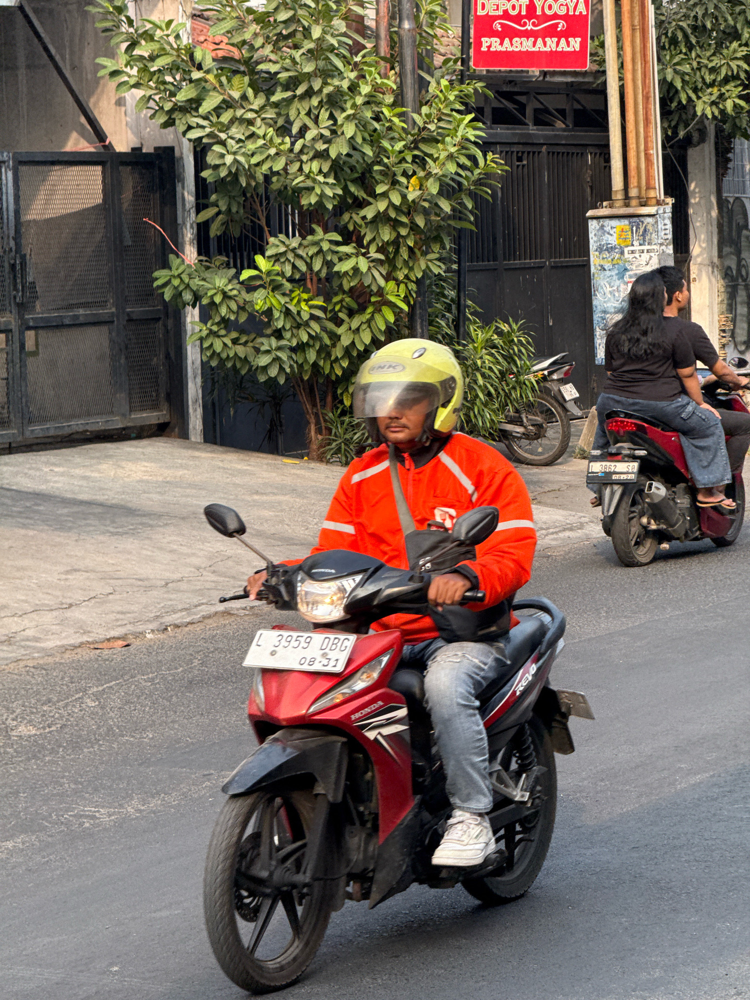 In Surabaya, Indonesia, a person wearing an orange jacket and a yellow helmet is riding a red motorcycle with a license plate that reads "L 3959 DBG 08-31". The motorcycle is on a paved street, and the person is looking forward. In the background, another person is riding a motorcycle with a black shirt and blue jeans. There are trees and buildings on the side of the street. A red sign with "DEPOT YOGYA PRASMANAN" is visible on one of the buildings. The sun is shining, casting shadows on the ground. The scene appears to be a typical street view in a city.