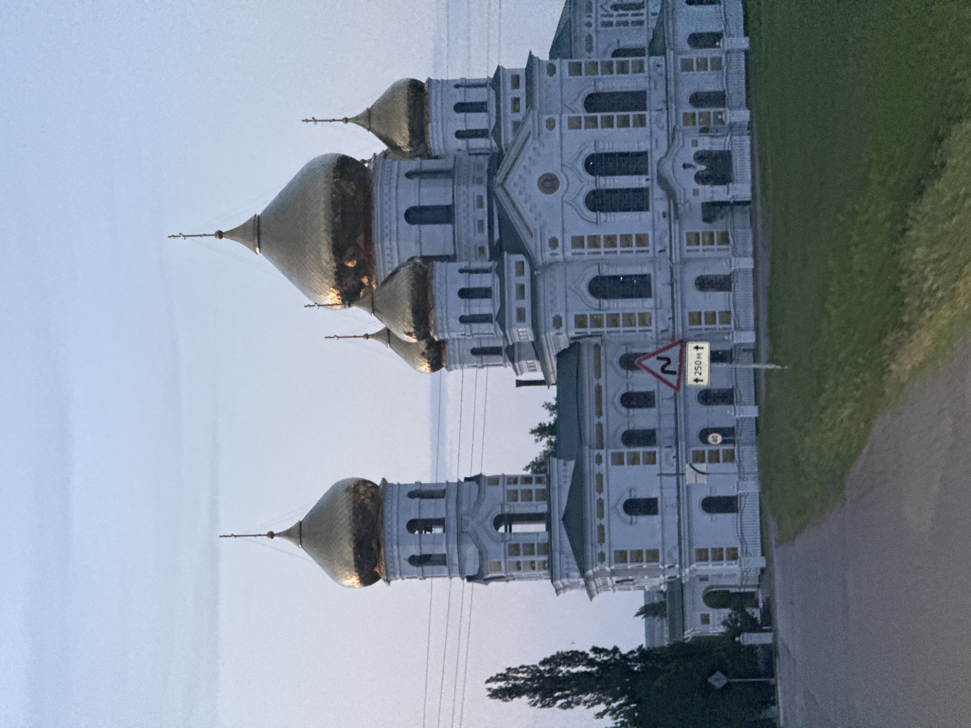 The image shows the Ascension Cathedral in Novodonetskaya, Russia. It is a white building with golden domes. A road runs along the side of the cathedral, with a grass field on the right. A sign warning about a turn is visible. Power lines are also visible in the sky. The sky is light blue, suggesting either dawn or dusk. Trees are visible at the bottom of the picture.
