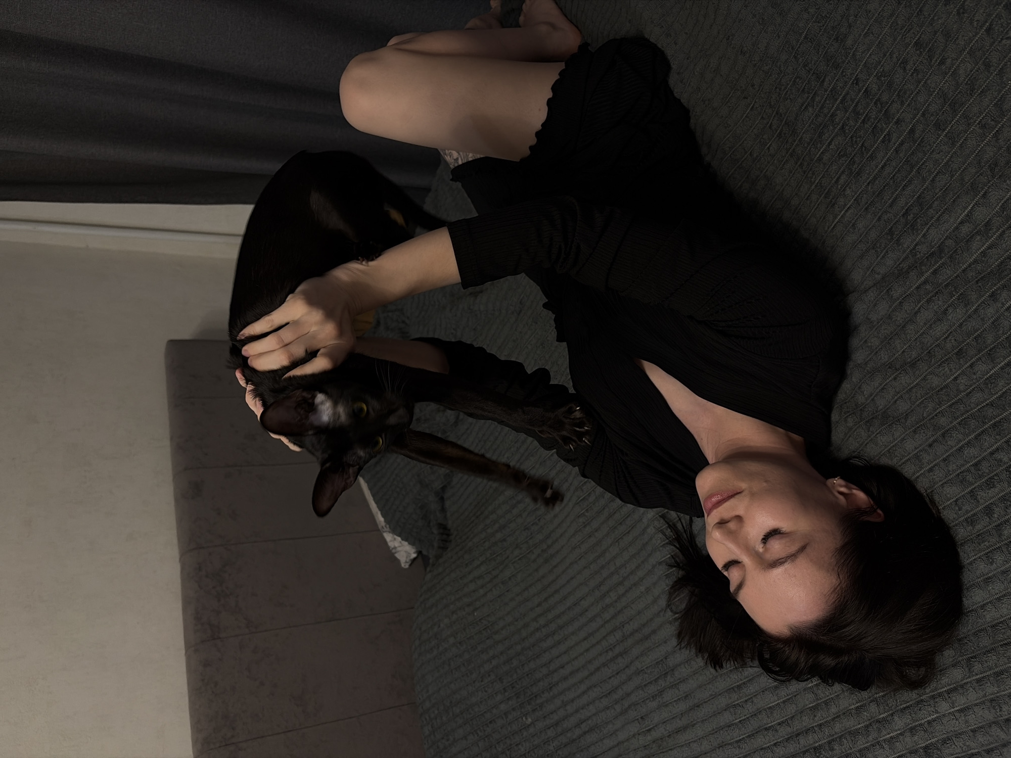 A woman in a dark dress lies on a textured gray bedspread, petting a black cat. The cat is oriented towards the left side of the frame. The woman's face is partially visible, with her eyes closed. The background includes a gray wall or curtain. The location is Chaikovsky, Russia.