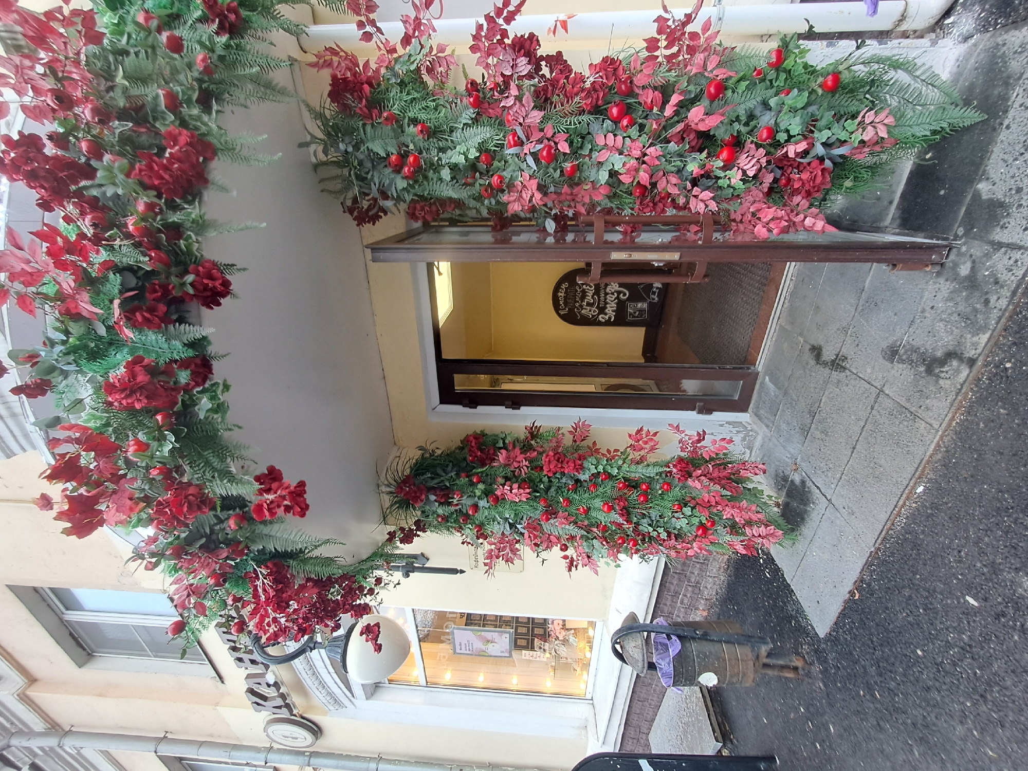 An eye-level outdoor shot, taken in Elektrostal, Russia, shows the entrance to a building decorated with red and green floral arrangements. The entrance features a doorway that leads into an interior space with a sign hanging in the background. The doorway is surrounded by a lavish wreath-like arrangement of red flowers and green foliage, extending over the entrance and down the sides. The building facade is painted in a light yellow color. A street lamp is visible on the left side of the frame. The foreground shows a section of pavement or sidewalk leading to the entrance. The shot conveys a festive and welcoming atmosphere, likely marking a special event or seasonal decoration.