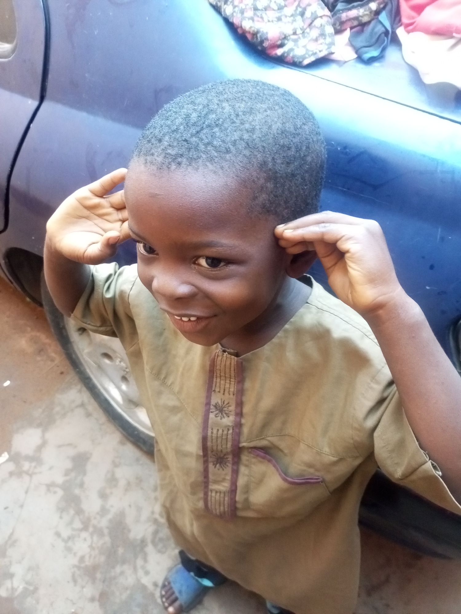 A young boy, approximately 6-8 years old, stands outside in what appears to be a dusty, urban environment. He has dark skin and closely cropped hair. The boy is wearing a light brown, long-sleeved garment with decorative trim on the chest and a small pocket on the right side. His right hand is raised, and he is touching his right ear. His left hand is also near his ear, possibly adjusting it or covering it. He is smiling slightly, with his mouth open. The boy is standing next to the rear wheel of a dark blue vehicle. In the background, the vehicle's body panel and some folded cloth items are visible. The ground is a mix of dirt and concrete. The boy is wearing blue sandals. The scene is illuminated by natural light, suggesting it is daytime. The location is identified as Tempure, Nigeria.
