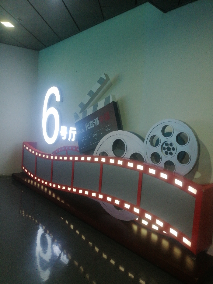 An indoor scene in Fengtai District, China, features a cinema-themed decorative display. Centrally positioned against a light blue-green wall is an illuminated white numeral "6" followed by the Chinese characters "号厅" (hào tīng), indicating "Hall 6." To the right and slightly behind, a brown-colored, stylized film clapperboard is visible, bearing the text "PROD: 光影看" (Guāng Yǐng Kàn) and fields for "SCENE," "TAKE," and "ROLL." Behind the clapperboard are two large, white, circular film reels. In the foreground, a curved, three-dimensional representation of a film strip with a red frame and grey sections exhibits numerous small, glowing white rectangular lights along its edges, simulating perforations. This entire display rests on a dark base. The dark, glossy floor below reflects the illuminated display elements. Overhead, a dark, paneled ceiling is partially visible, with one square light fixture reflecting on the floor. No individuals are present, and no actions are occurring.