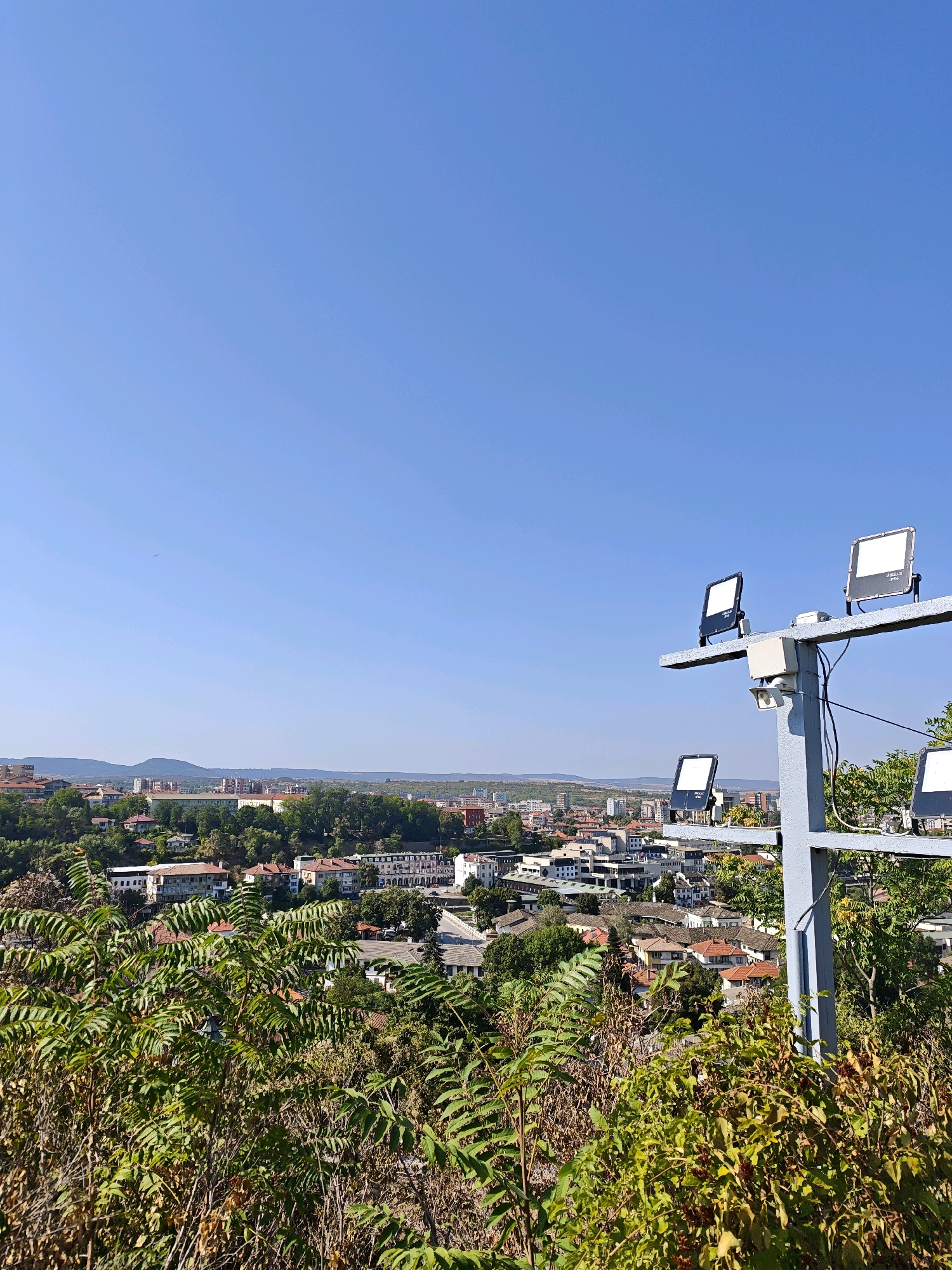 A high-angle, eye-level, medium shot shows a clear blue sky above a cityscape in Lovech, Bulgaria. In the foreground, a metal pole with four floodlights and a security camera is mounted. The floodlights are white and rectangular, with one pointing left, one right, and two pointing forward. The security camera is small and white, positioned below one of the floodlights. Behind the pole, a dense cluster of green foliage obscures the lower part of the cityscape, with some dry, brown brush visible at the bottom of the frame. The city below features a mix of buildings with red-tiled roofs and larger, modern structures. In the distance, a range of low hills and mountains is visible under the clear sky.
