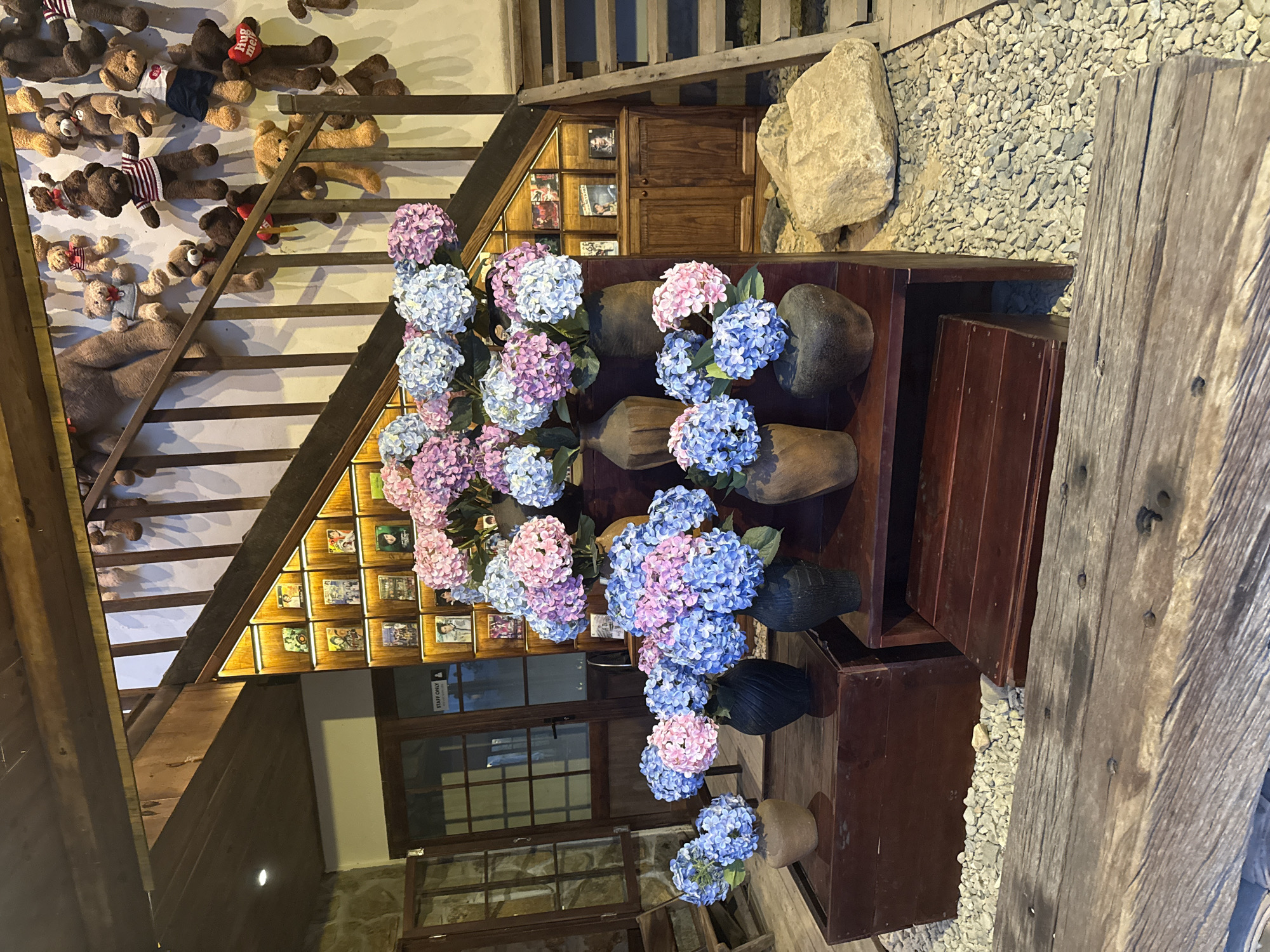 The image depicts an indoor scene, possibly a cafe or a shop, featuring a wooden staircase and an array of decorative items. Several vases filled with pink and blue hydrangeas are prominently displayed on wooden pedestals and a shelf. Above the staircase, a collection of stuffed teddy bears are hanging on the wall. On the wall beneath the staircase, there are shelves filled with what appear to be framed photos or album covers. In the background, a wooden cabinet with doors is visible. The floor is covered with gravel and large rocks. The overall ambiance suggests a whimsical and cozy setting. The specific location in Nam Nha Trang Ward, Vietnam, is not visually identifiable from the provided image.