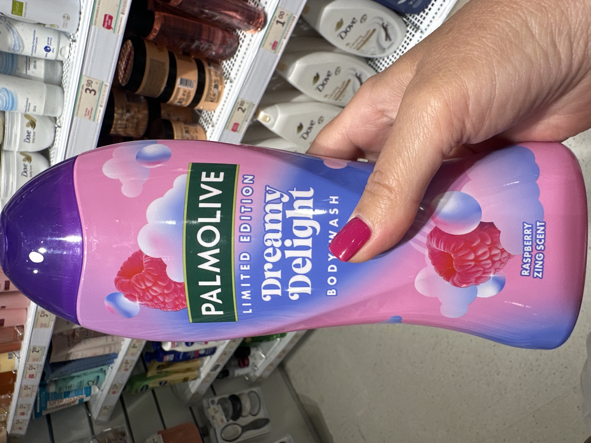 An outdoor, eye-level, medium shot shows a hand holding a Palmolive Dreamy Delight body wash bottle. The bottle is pink and purple with graphics of raspberries and clouds. The text on the bottle reads "PALMOLIVE LIMITED EDITION Dreamy Delight BODY WASH RASPBERRY ZING SCENT." The background appears to be a retail store aisle with shelves of various personal care products, including Dove products. The lighting is bright and even.
