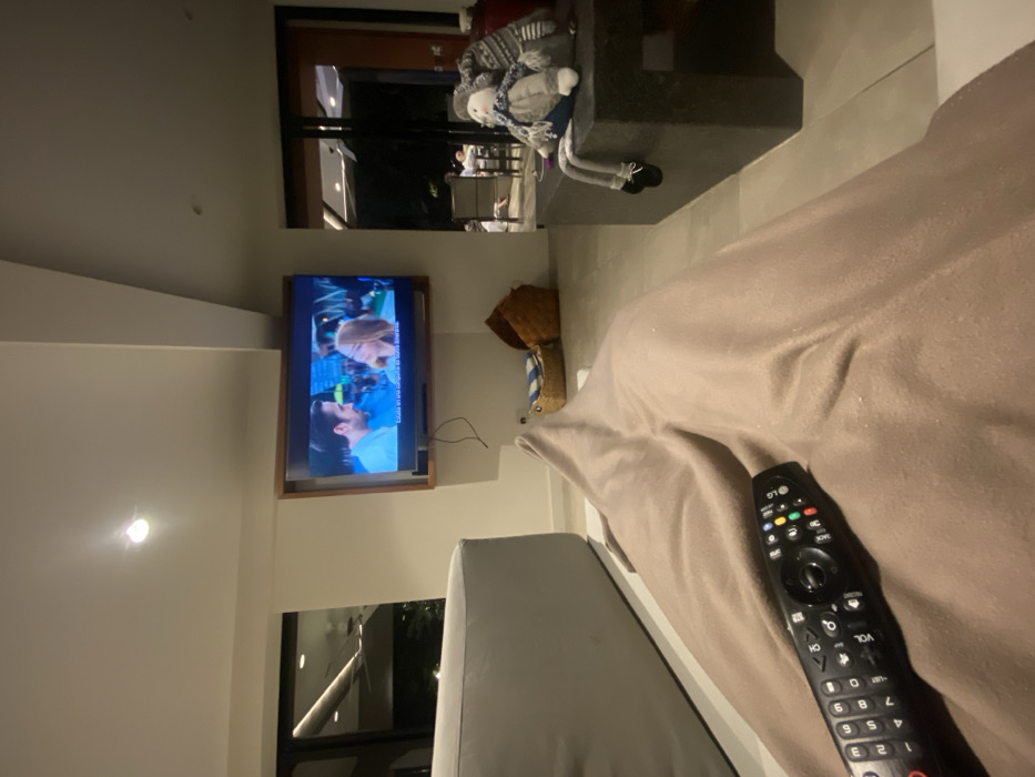 An indoor view shows a television mounted on a white wall. The TV is displaying a scene with two individuals in close proximity. Next to the TV is a decorative straw basket. A tan-colored soft chair or bed is in the foreground, with a black LG remote control lying on its surface. A plush doll is placed on a nearby surface in the background. The shot is taken from a slightly elevated position, providing a partial view of a window.