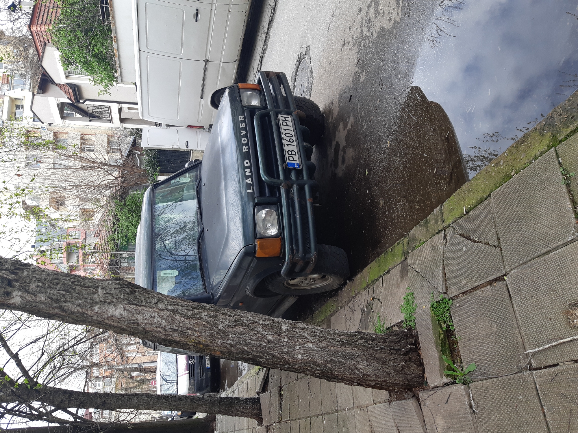 The image depicts a Land Rover parked near a curb in Plovdiv, Bulgaria. A tree trunk is positioned between the sidewalk and the front of the vehicle. A puddle of water reflects the sky and nearby buildings. A white van is parked behind the Land Rover. The license plate number of the Land Rover is PB 1601 PH. The sidewalk is paved with patterned concrete blocks, and some are cracked. Green moss and small plants grow in the cracks. Buildings are visible in the background. Bare tree branches obscure some of the scene. A black vehicle with the license plate CB 3688 is partially visible behind the tree.