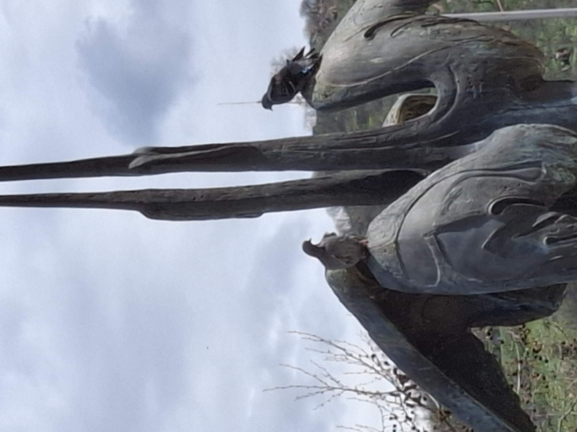 A low-angle, close-up shot shows a bronze statue in Plovdiv, Bulgaria, likely outdoors due to visible vegetation and a cloudy sky. The statue depicts a figure or figures rendered in an abstract, metallic style. Two pigeons are perched on the statue. One is on its head and one is on its wings.