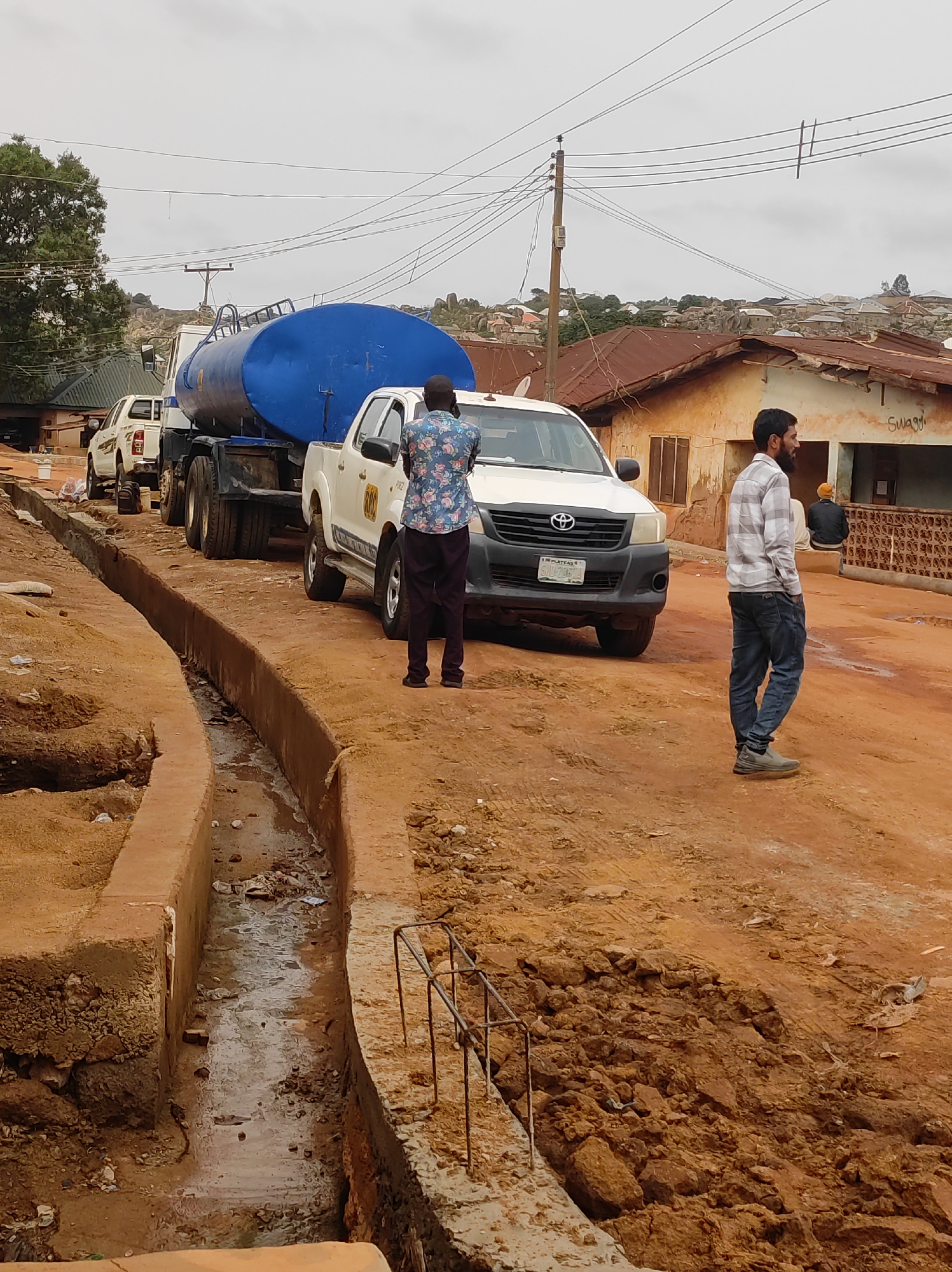 The image depicts a street scene in what appears to be the city of Jos, Nigeria. A white Toyota Hilux pickup truck towing a blue water tanker is parked on an unpaved red dirt road. Two men are standing near the vehicle. One man, wearing a patterned shirt and dark trousers, is facing the truck. The other man, wearing a plaid shirt and jeans, is standing near a building with a partially visible sign. In the foreground, a recently constructed concrete drainage ditch runs along the road. The background features houses with corrugated iron roofs and a few trees under an overcast sky. In the background, powerlines are seen overhead. A second white pickup truck is parked in the background as well. The license plate of the car states "Plateau - State of Peace".