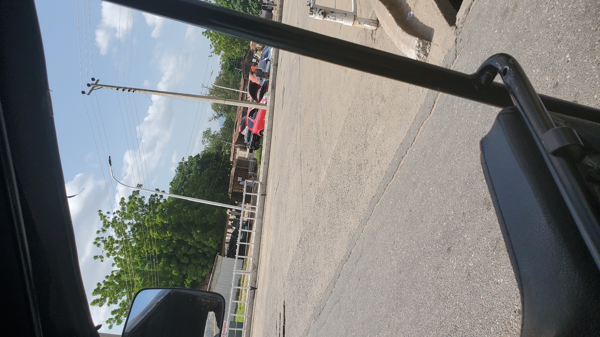 The image captures a street scene in Jalingo, Nigeria, viewed from inside a vehicle, likely a tricycle or similar open-air transport. In the foreground, a black padded seat and black metal bars of the vehicle's frame are visible. The road surface is asphalt with visible cracks and patches.
Beyond the road, a white metal railing separates the street from a sidewalk or verge. Several buildings, appearing to be shops or small businesses with corrugated metal roofs, line the area behind the railing. A red car is parked with its hood open, and a person is standing next to it, possibly attending to the vehicle. Adjacent to it, a dark-colored car is also visible. Further down the street, a few more people are seen standing or walking.
The environment is characterized by a bright, sunny day with a partly cloudy sky. Green trees are abundant, suggesting a lush area. Utility poles with electrical wires traverse the sky, and a modern street lamp is also present. The overall impression is of a typical urban or peri-urban setting in Jalingo.
