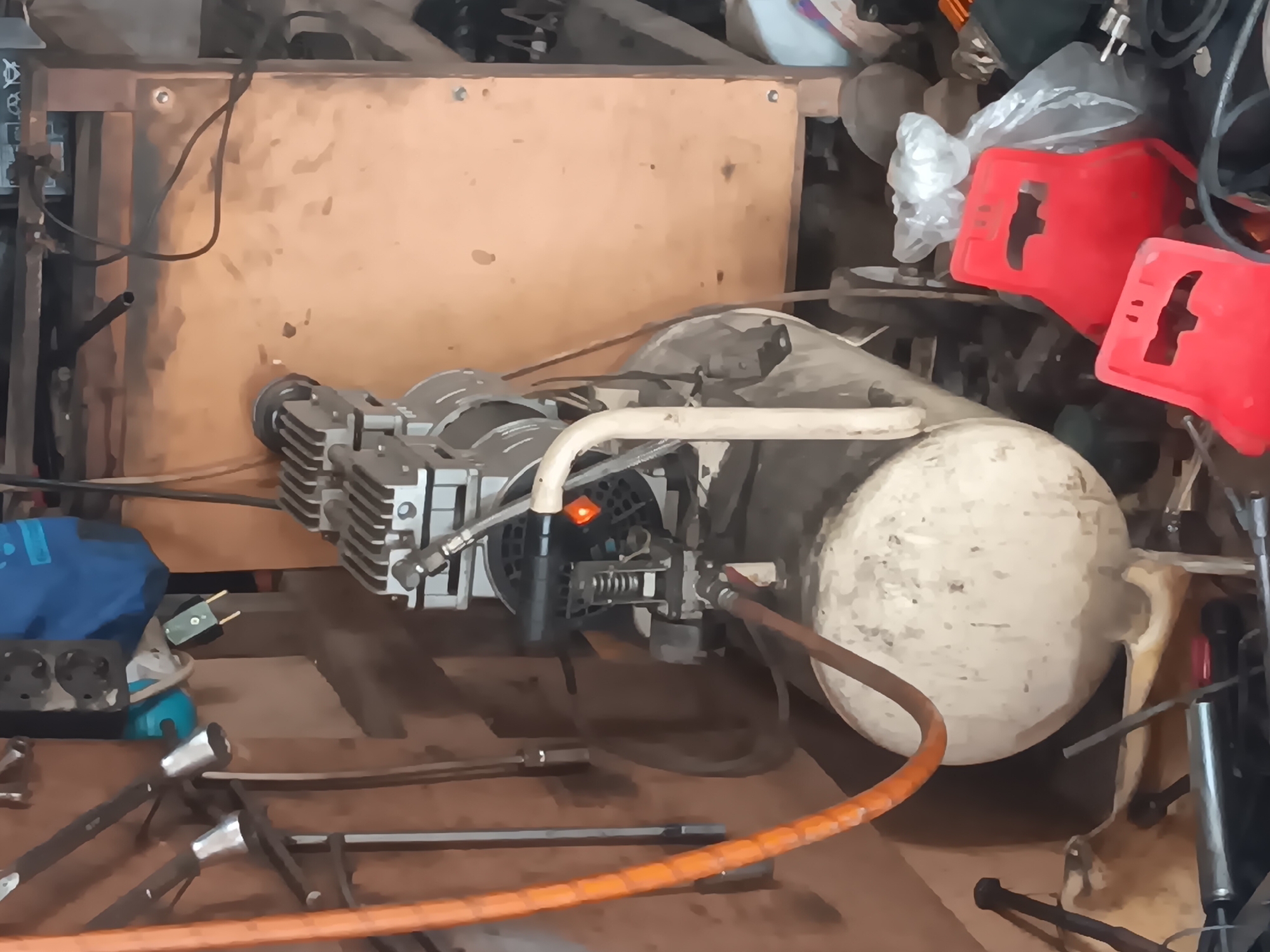 The image depicts a cluttered workshop or storage area. In the center, a portable air compressor with a large, off-white tank and a silver motor assembly is the primary subject. An orange hose is coiled and attached to the compressor. To the right of the compressor, two bright red plastic seats are stacked, partially obscuring a clear plastic bag containing various items. The background consists of wooden panels and shelves filled with tools and equipment, including what appear to be wrenches and other metal objects hanging on the left wall. The scene is dimly lit, suggesting an indoor environment. There are no people visible in the image. The overall impression is one of disarray and a functional, working space. The provided coordinates indicate a location in Batursari, Indonesia.