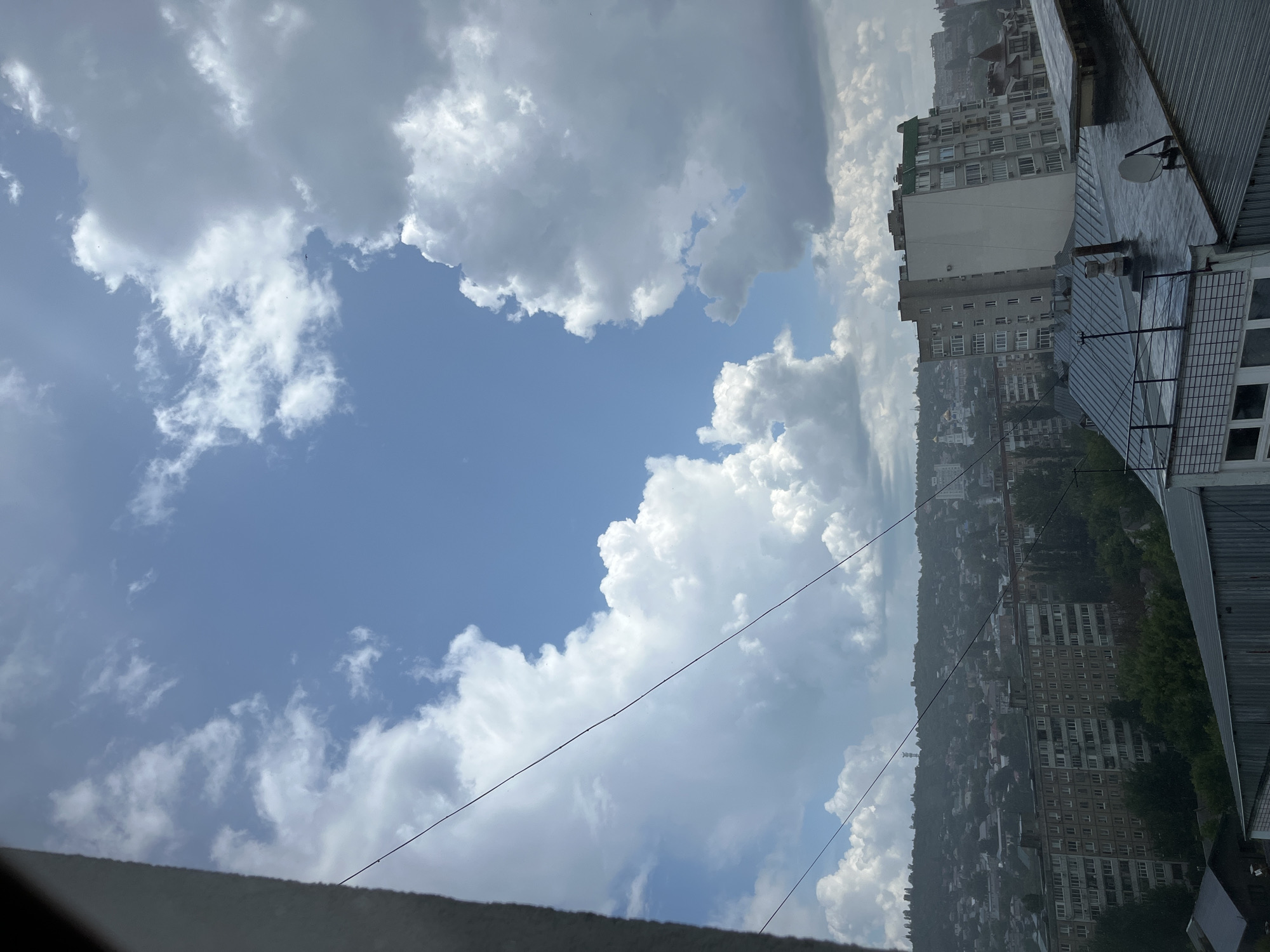 A wide-angle shot captures a cloudy sky over a city, likely Saratov, Russia, judging by the architecture and topography. The sky dominates the upper half of the frame, filled with large, puffy white clouds against a bright blue background. A thin, dark power line stretches diagonally across the sky. Below the clouds, a dense urban landscape unfolds. Numerous apartment buildings, mostly of Soviet-era design, are visible, with a hilly background covered in trees and smaller structures. A prominent, modern apartment building with a green roof stands out among the older buildings on the right side of the frame. The foreground reveals the metallic roof of a building, wet from rain, with visible pipes and a satellite dish. No people are visible in the image. The overall impression is of a typical cloudy day in a densely populated city.