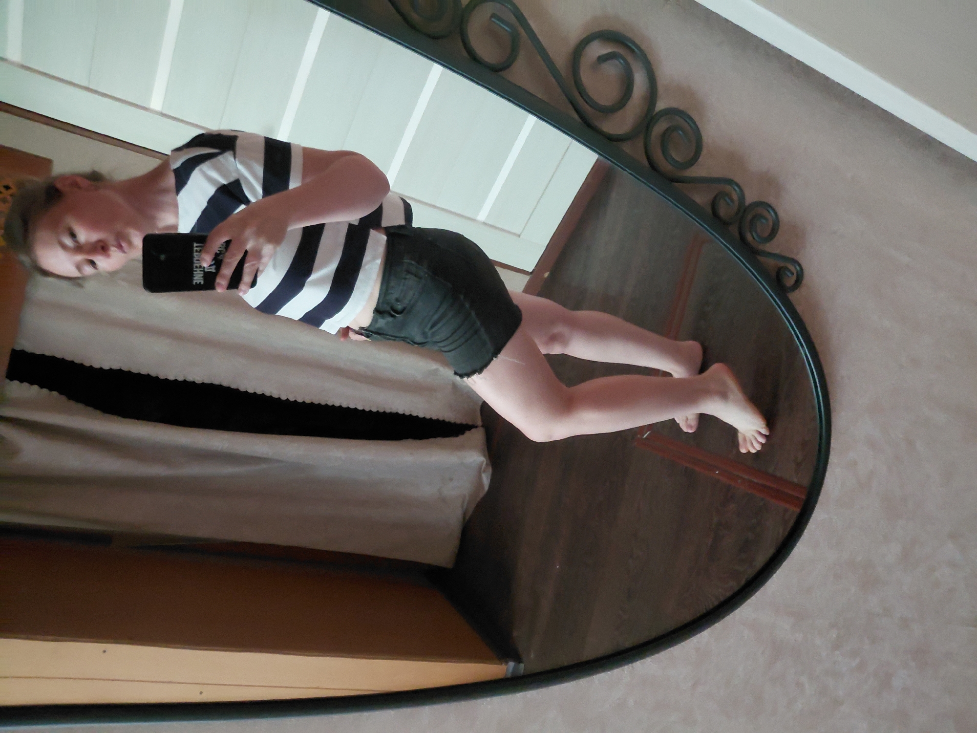 A woman stands facing a mirror, taking a selfie. She has light skin and light brown hair pulled back. She is wearing a black and white striped t-shirt and dark denim shorts. Her right hand is holding a black smartphone, and her left hand is resting on her hip. The background shows a curtain, a wooden door, and a wall with textured wallpaper. The floor is wood. The mirror has an ornate dark metal frame. The image does not provide any specific details to determine the location within the city of Novosibirsk, Russia.