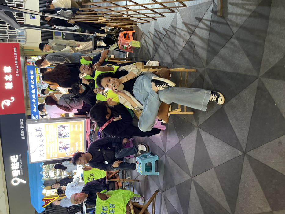 The image shows a street scene in Hang Zhou Shi, China, at the coordinates 30.264 latitude, 120.155 longitude. There are several people in the scene, including a young woman wearing blue jeans and a black shirt sitting on a wooden chair, a young man seated beside her wearing a black shirt, and several other people standing in the background, some of whom are wearing yellow shirts. There are also various objects in the scene, including a street sign, a metal barrier, a folding chair, and a variety of other items. The environment is a city street with a tiled floor and a street sign that reads "No. Nine Shoes Club." The people are interacting with each other. The image was captured from a slightly elevated angle, providing a clear view of the scene.