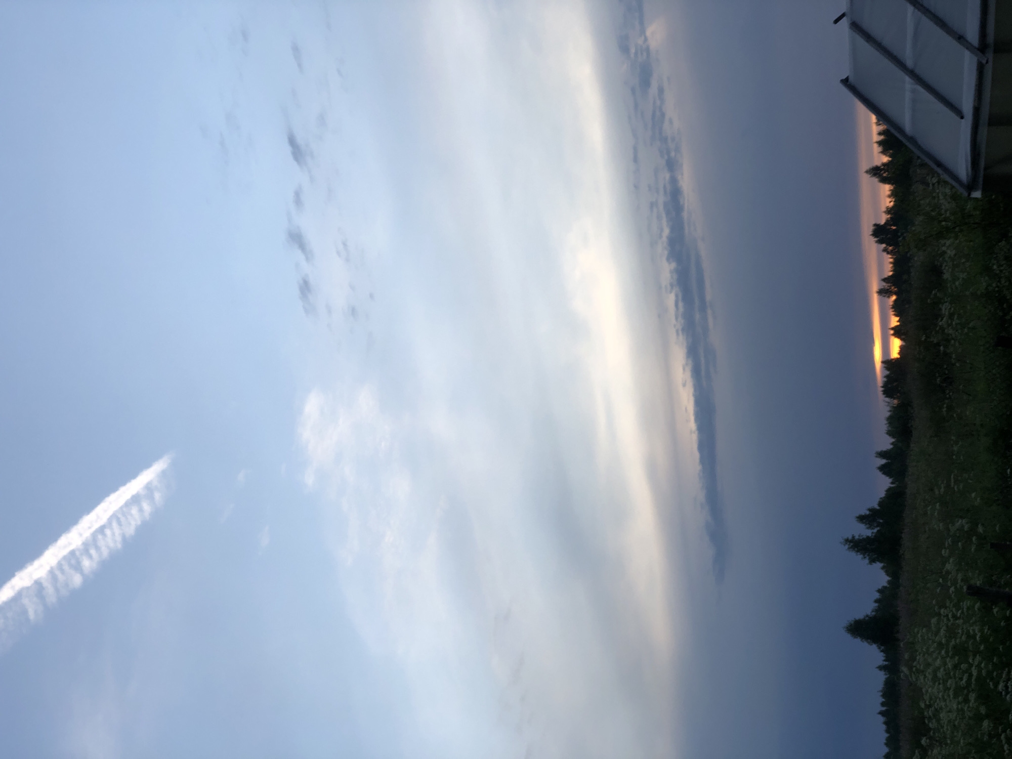 This is a vertical, eye-level, full shot of a dusky sky over a field and forest line with a greenhouse in the foreground. The sky is a pale blue with a few wispy clouds and a faint contrail from an airplane. The sun is setting on the horizon, casting a warm orange glow. The field is covered in tall grass and wildflowers, and the forest line is dark and silhouetted against the sky. A greenhouse with a translucent roof is visible on the right side of the frame. There are no people or animals in the image. The scene appears to be in a rural area outside of Uriupino, Russia.