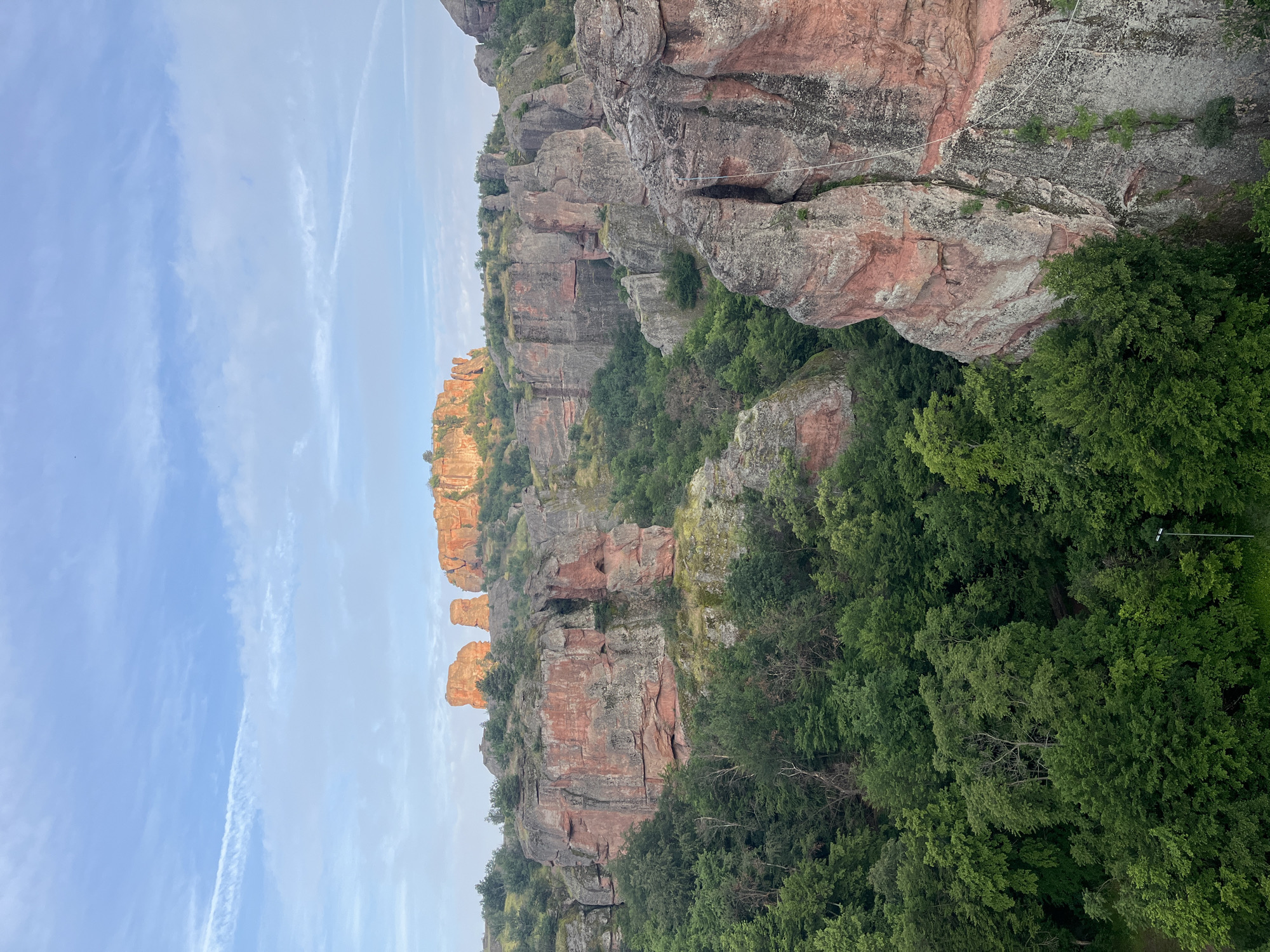 An outdoor, high-angle panoramic photograph captures a sprawling natural landscape featuring distinctive red and grey sandstone rock formations. Although the specific city and location are designated as Unknown, Unknown, the geological features strongly resemble the Belogradchik Rocks in Bulgaria. The scene is illuminated by the warm, directional light of either early morning or late afternoon. The sky occupies the upper third, a clear light blue with scattered white, wispy clouds and faint contrails. In the background, several massive, rounded rock pillars and formations glow with intense orange and red hues where they catch direct sunlight. The midground comprises a complex series of towering cliffs, deep ravines, and eroded rock walls, primarily reddish-brown with grey and white striations, interspersed with dense pockets of dark green deciduous trees and shrubs clinging to the slopes and crevices. The foreground is a rich tapestry of vibrant green trees, forming a dense canopy that partially obscures the bases of the rock formations. A white slackline or rope is visible stretched horizontally across two prominent rock faces in the lower right section of the image. A single modern streetlamp with a grey pole stands upright amidst the trees in the far bottom-right. No people are present in the scene.