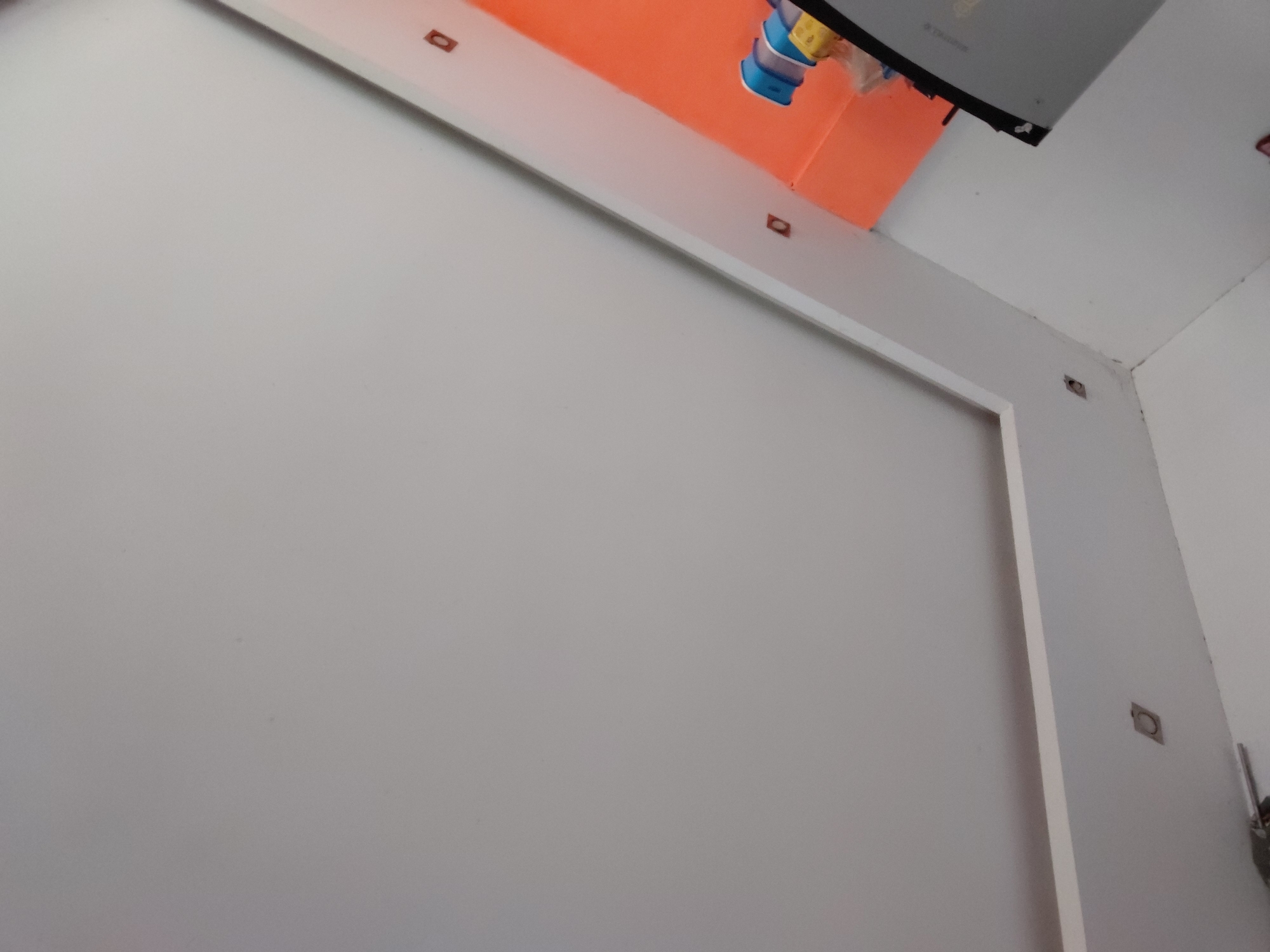The image displays an indoor setting, likely a room under construction or renovation in Santa Ana, Venezuela. The scene is dominated by light gray/white walls and a white ceiling. A large, L-shaped white architectural trim frames a section of the main wall, suggesting a recessed area or decorative panel. Above this trim, a vibrant orange structure, possibly a suspended ceiling element or a large fixture, is prominent.
From the area above the orange structure, several blue and yellow cables hang downwards, alongside an inverted gray monitor or screen. Faint, inverted text on the back of this screen appears to read "VIZIO". Small, dark, rectangular cutouts or fixtures are embedded at intervals along the top edge of the white trim, and one similar cutout is visible in the upper right corner where the wall meets the ceiling. On the right-hand wall, a raw, uncovered cutout for an electrical box is visible, further indicating an unfinished state. No people are present, and the lighting appears to be even, typical of indoor artificial light or diffused natural light.