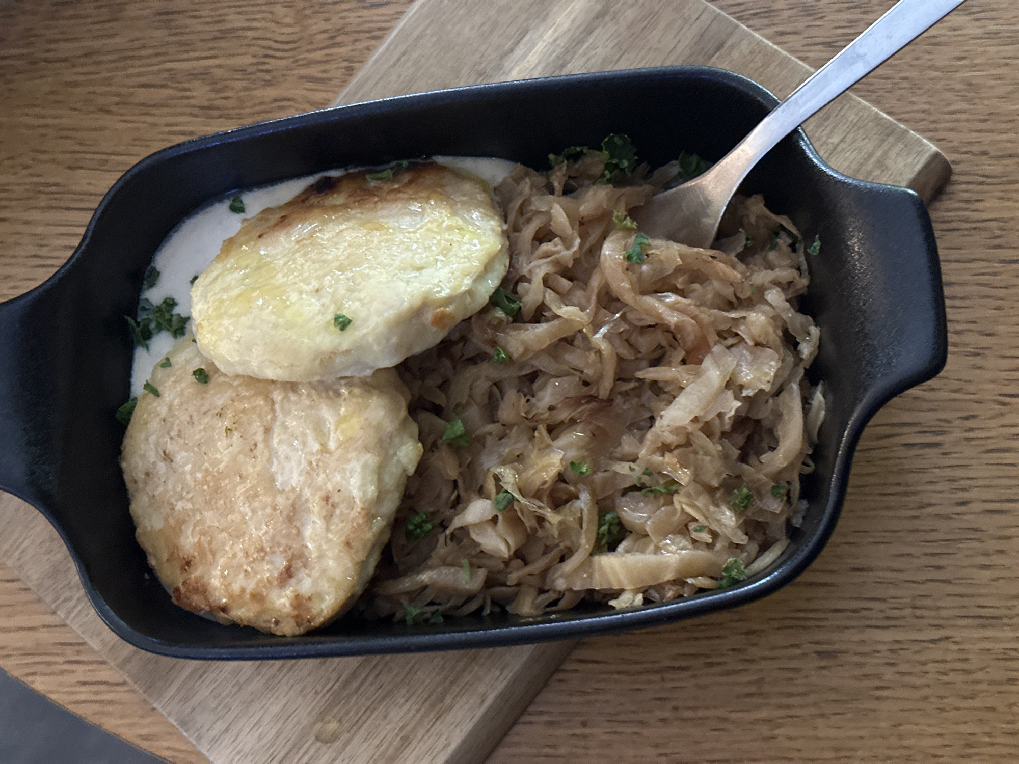 An overhead shot displays a hearty meal served in a black rectangular cast-iron dish, resting on a light brown wooden surface, possibly a serving board or table. The dish contains two prominent, round, pan-fried items, which appear golden brown and slightly crisp on top, possibly potato pancakes, dumplings, or patties. These are partially submerged in a creamy, white sauce, which also serves as a base.
To the right of these items, the dish is filled with a large portion of finely shredded and sautéed vegetables, likely braised cabbage or sauerkraut, exhibiting a soft, golden-brown hue. Scattered green specks, presumably chopped parsley or other herbs, are visible as a garnish over both the creamy sauce and the sautéed cabbage, adding a touch of freshness. A silver fork is partially visible, resting in the dish among the shredded vegetables, suggesting the meal is ready to be eaten. The overall presentation is rustic and appealing, indicative of traditional, comforting cuisine. There is no visible text in the image.
