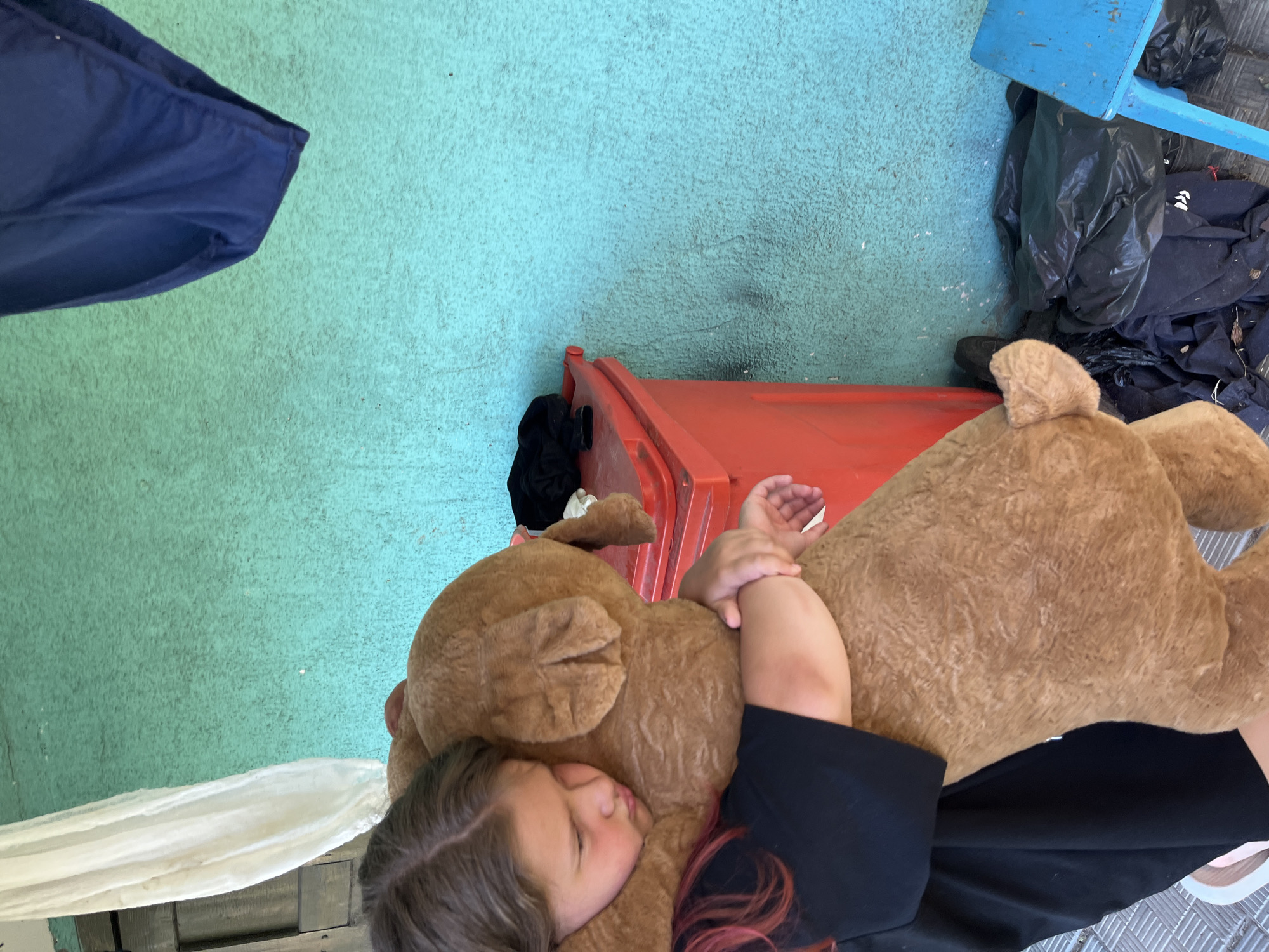 In Navahrudak, Belarus, a young girl with reddish-brown hair pulled back and wearing a black t-shirt hugs a large, light brown stuffed bear. She is positioned on the left side of the frame, her face turned towards the bear, with her eyes closed and lips pursed. Her arms are wrapped tightly around the bear, holding it close. The girl is standing next to a bright red trash can, which is filled with dark-colored items, including black clothing on top. To the right of the trash can, a pile of black garbage bags is visible on the ground. The background features a textured, turquoise-colored wall. A light-colored curtain hangs on the left side of the wall, and dark blue undergarments are suspended from the top right of the frame. A small blue stool is situated on the far right of the image. The ground beneath the girl and the objects is a light gray, textured surface.