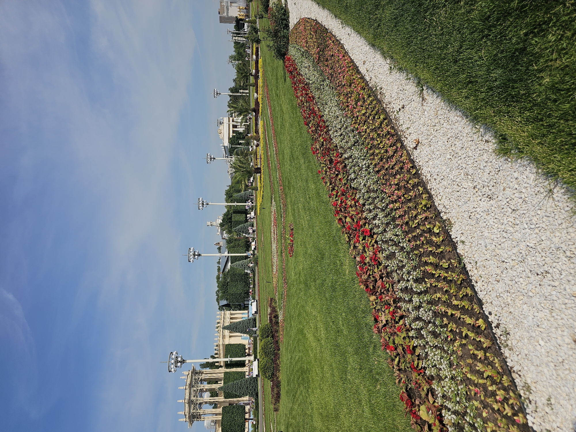This image captures a wide-angle view of the Exhibition of Achievements of National Economy (VDNKh) in Moscow, Russia, under a clear blue sky with minimal wispy clouds. The foreground features a meticulously maintained landscape. On the right, a strip of vibrant green grass runs alongside a curved path of white gravel. To the left of the gravel path, a long, sinuous flowerbed displays distinct bands of plants: deep red blooms, white flowers, and light green/pinkish foliage plants arranged in linear rows. Beyond this, a vast expanse of manicured green lawn extends into the midground, dotted with circular and oval-shaped flowerbeds containing various colorful plantings and small evergreen shrubs.
In the midground, several tall light poles, each topped with multiple spherical luminaires, are spaced throughout the park. Manicured trees, including conical evergreens and box-shaped hedges, line the pathways and frame the background architecture. Several palm trees are also visible within the distant flowerbeds. Small figures of people are discernible walking on paths in the distance, and several small white utility or service vehicles are present near the buildings.
The background is dominated by the grand, ornate architectural structures characteristic of VDNKh. On the left, an elaborate, light-colored, open-air pavilion or gazebo with classical columns and intricate decorative elements stands prominently. Further into the background, several large, cream-colored exhibition pavilions with classical facades, arches, and columns are visible. To the far right, a partial view of another grand structure features what appears to be a golden element, possibly part of a fountain or monument. The scene is brightly illuminated by direct sunlight, casting visible shadows.
