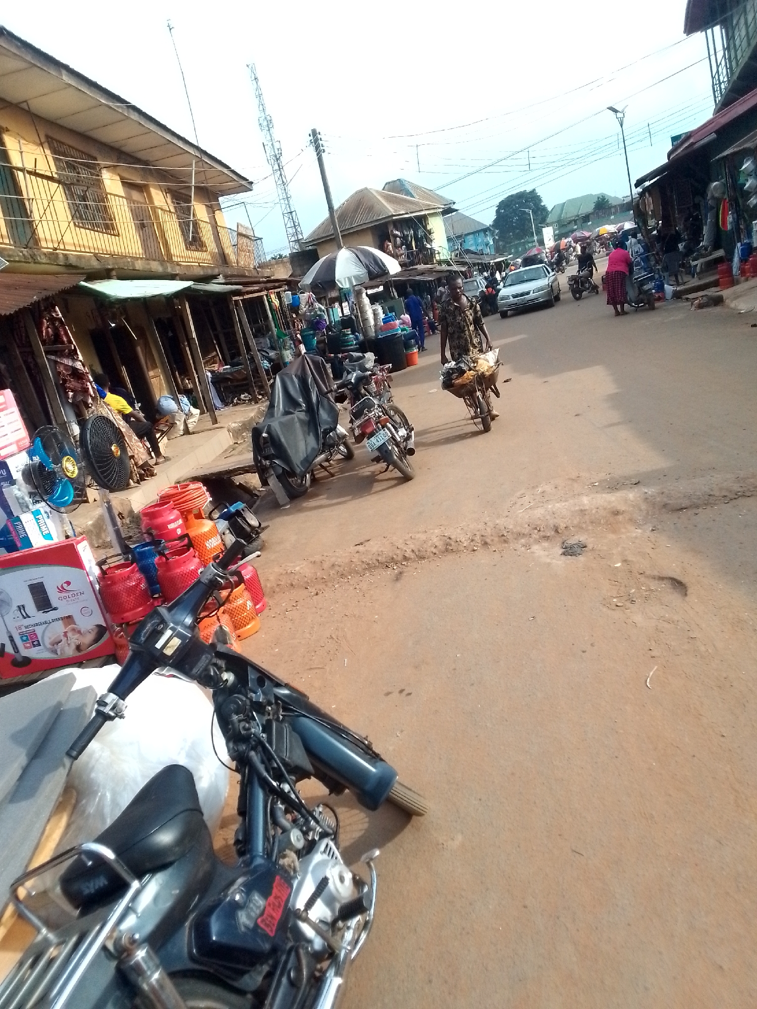 The image depicts a street scene in Orlu, Nigeria. The road is paved but shows signs of disrepair with potholes. Several buildings line the street, some of which appear to be shops with awnings. People are walking along the street, and several motorcycles are parked or in motion. A car is visible further down the street. There are goods displayed for sale, including gas cylinders, fans, and other items. The scene gives the impression of a busy commercial area in Orlu.