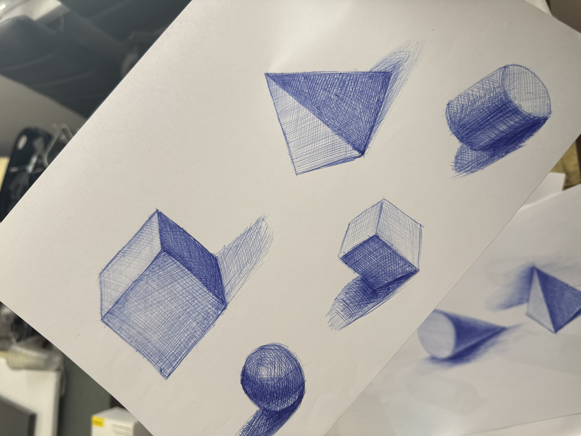 A white sheet of paper features pen drawings of geometric shapes, including a pyramid, cylinder, cube, and sphere. Each shape is shaded to create a three-dimensional effect, and each has a cast shadow. A second sheet of paper with similar drawings is partially visible in the lower right corner. The photograph was taken in an indoor environment with artificial lighting. The location is unknown and cannot be confirmed as Sofia, Bulgaria from the given image.