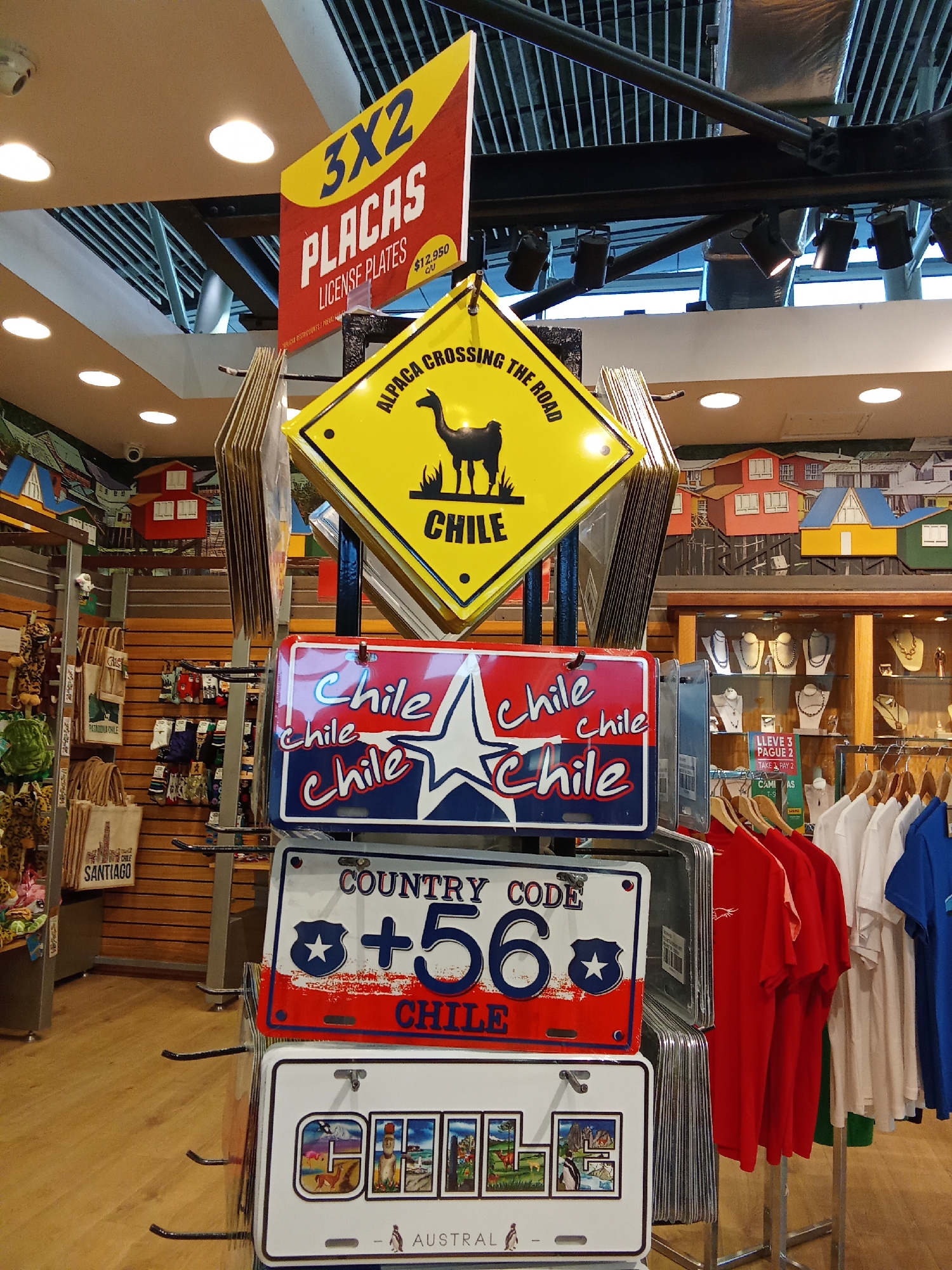 This is a medium shot, eye-level, indoor photograph of a retail display featuring novelty license plates and merchandise. The display is set up with multiple tiers of hanging items. In the foreground, there are several novelty license plates. The top plate in the stack is red, white, and blue, with "CHILE" repeated in cursive and block letters, along with a white star. Below it, another plate is predominantly white with "COUNTRY CODE +56" printed in bold blue letters. It also features a red and blue background with a white star at the top left. The bottom-most visible plate is white with a montage of scenic images and text that reads "AUSTRAL" at the bottom.

Above these license plates, a yellow diamond-shaped sign is visible, with black text that reads "ALPACAS CROSSING THE ROAD" and a silhouette of an alpaca. Above that, a red and yellow rectangular sign states "3X2 PLACAS LICENSE PLATES" and displays a price of "$12,950 CL".

In the background, a wall mural depicts colorful houses on stilts. To the right, a glass display cabinet showcases necklaces and jewelry. Below the cabinet, a rack of t-shirts in red, white, and blue are hanging on hangers. Other merchandise, such as bags and socks with "CHILE" and "SANTIAGO" branding, are visible on shelves to the left. The overall environment appears to be a gift shop or souvenir store. The scene is well-lit, suggesting artificial lighting. There are no people visible in the frame. The location is identified as Pudahuel, Chile.