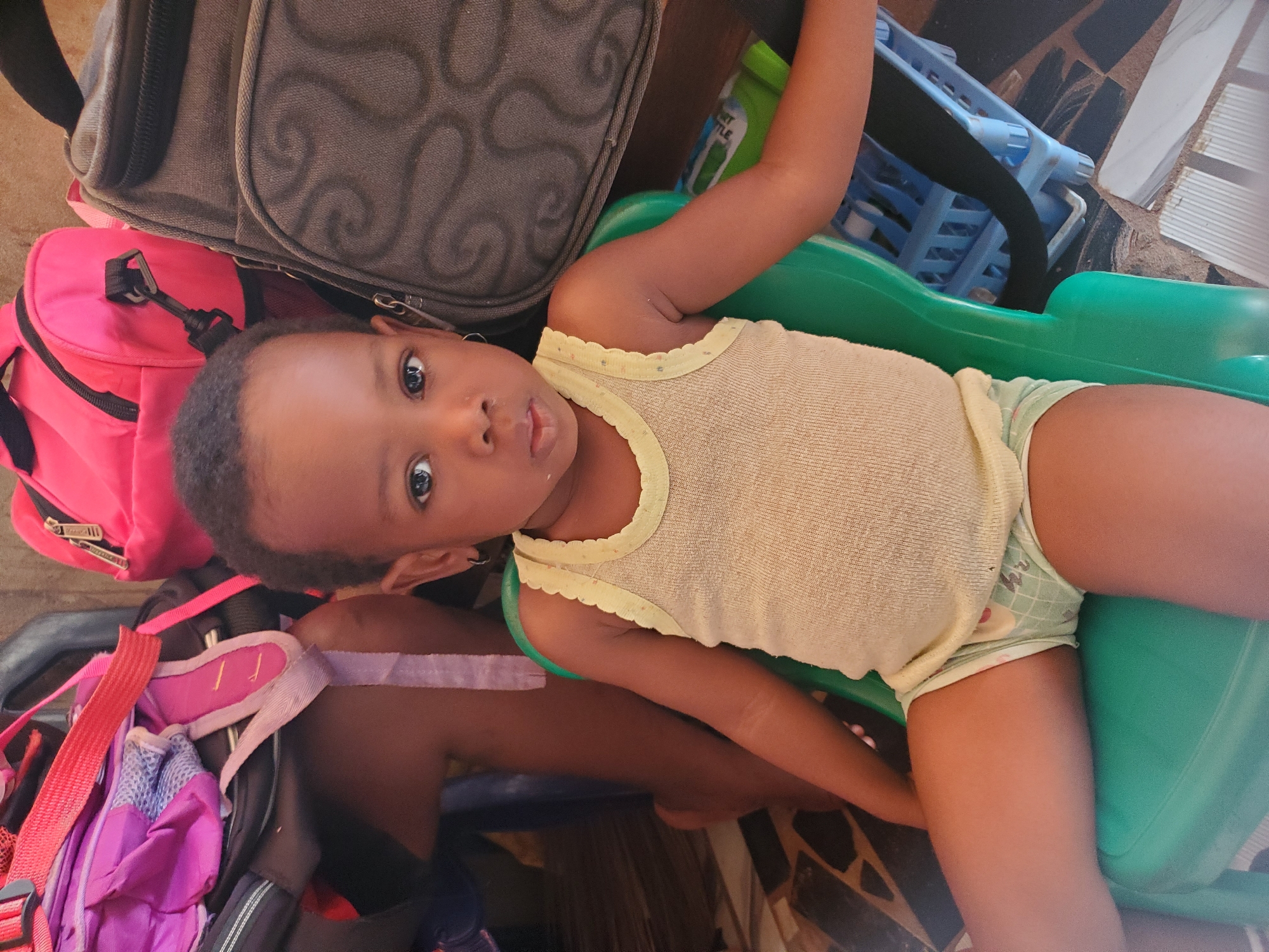 A child wearing a light yellow tank top and patterned diaper is seated in a green plastic child's chair. The chair is surrounded by luggage, including a gray suitcase, a pink backpack, and a purple backpack with pink straps. The background appears to be indoors, with a tiled floor visible. The location provided is Ukwu-Abwa, Nigeria.