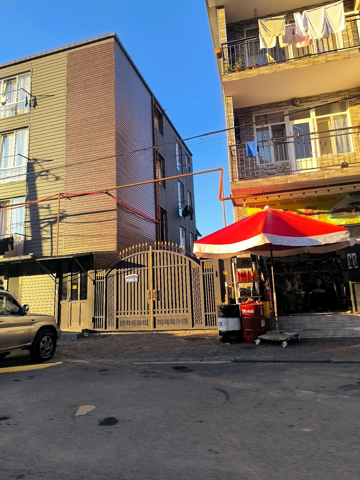 The image depicts a street scene in Salibauri, Georgia, under a clear blue sky. On the left, a multi-story building with beige and brown siding is visible, featuring several windows and balconies with laundry hanging out to dry. Adjacent to it, on the right, is another building with a yellow-tiled facade and balconies. Red pipes run along the exterior of both buildings.
In the foreground, a section of a paved road and sidewalk is present. A light-colored SUV is parked on the left side of the road. A large, ornate metal gate stands between the two buildings, leading to an entrance.
Beneath the yellow-tiled building, a storefront is visible, partially shaded by a large red and white umbrella. Several barrels, one labeled "Mobil," are placed in front of the storefront. Inside the store, two people are seated, appearing to be engaged in conversation. Various items, including bottles and shelves of products, are visible within the store. The overall impression is of a commercial street with residential elements, bathed in warm sunlight.